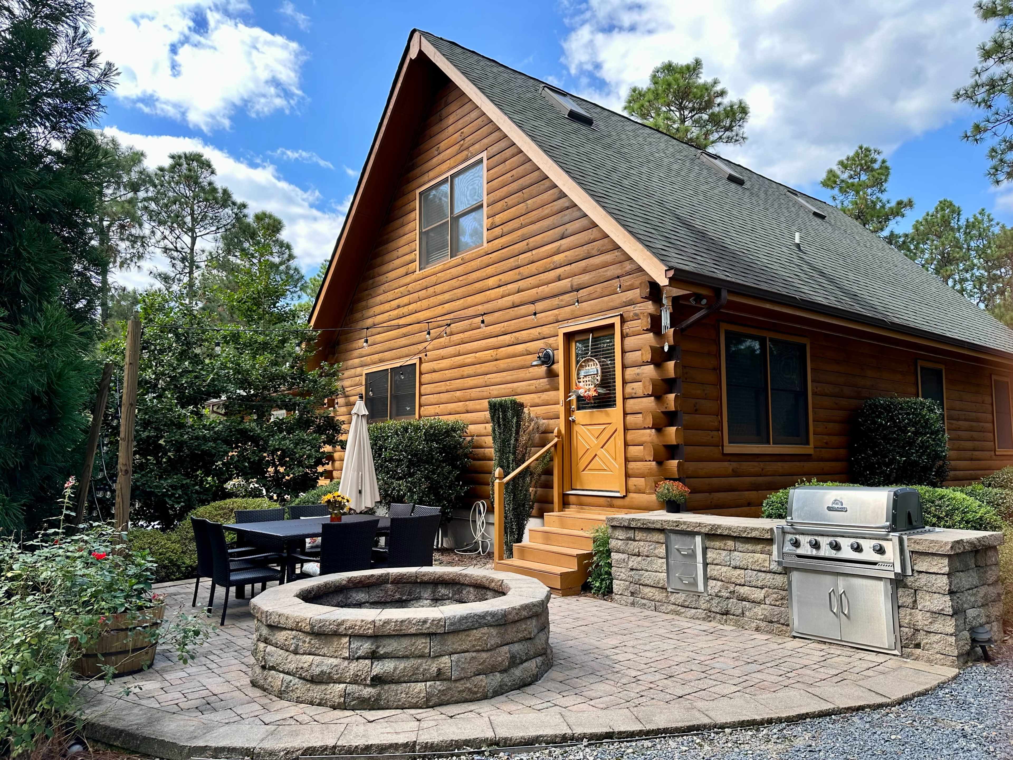 A log cabin with a stone patio features a fire pit, outdoor dining area, and a grill, surrounded by greenery and under a partly cloudy sky.