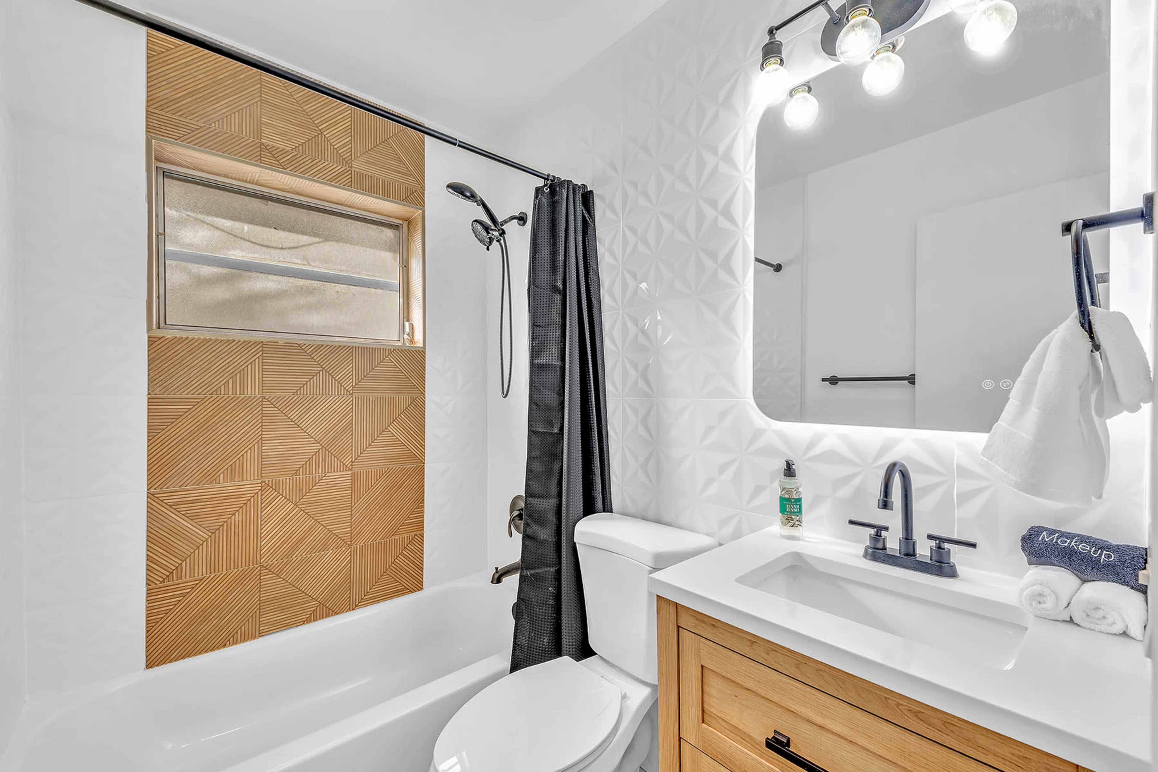 A modern bathroom featuring a bathtub, a wooden accent wall, a black shower curtain, a white countertop with a sink, and a round mirror with light fixtures above.