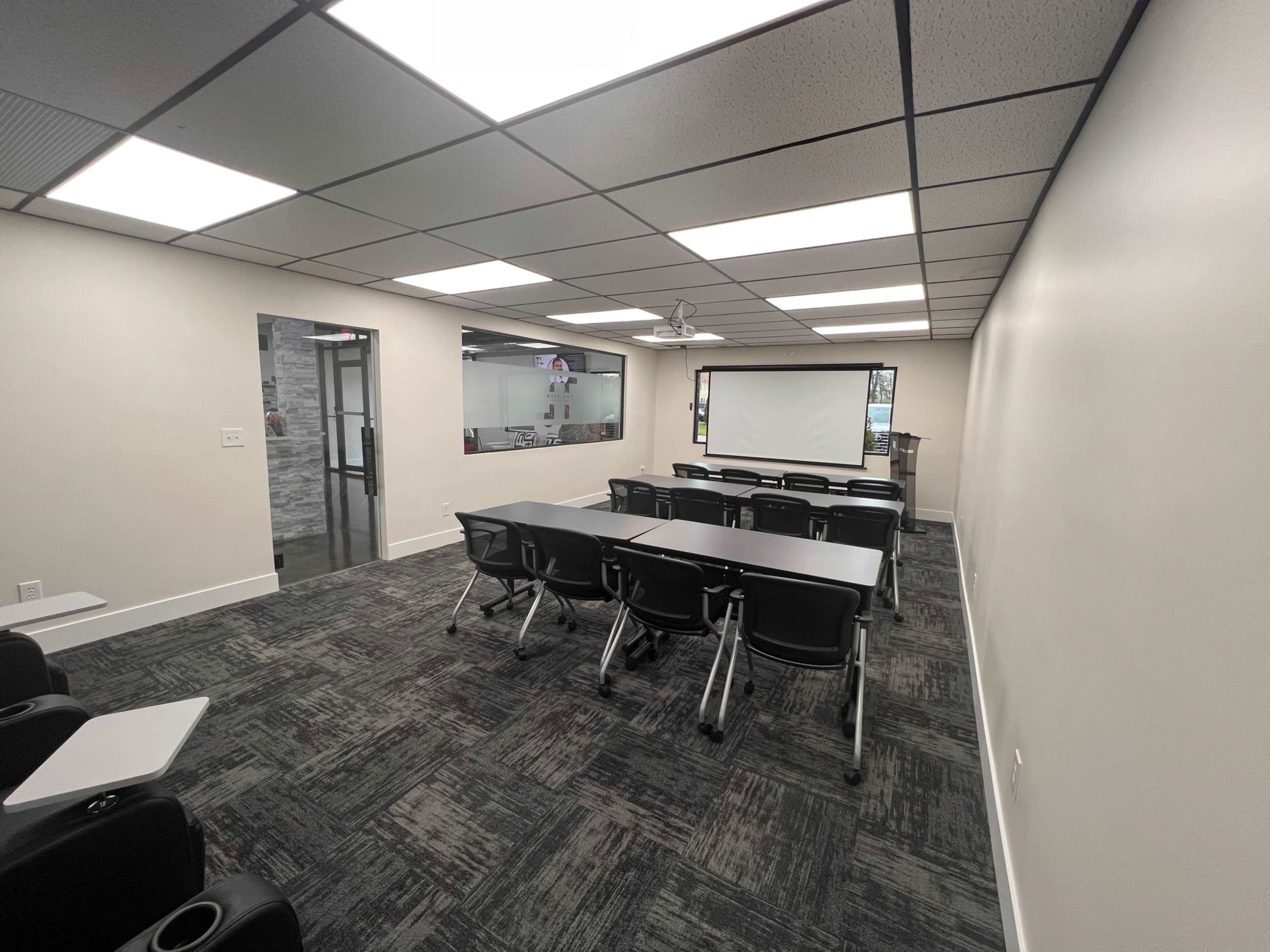 The image shows a modern classroom setup with rows of black chairs and tables, a projector screen at the front, and a glass-paneled door leading to another room.