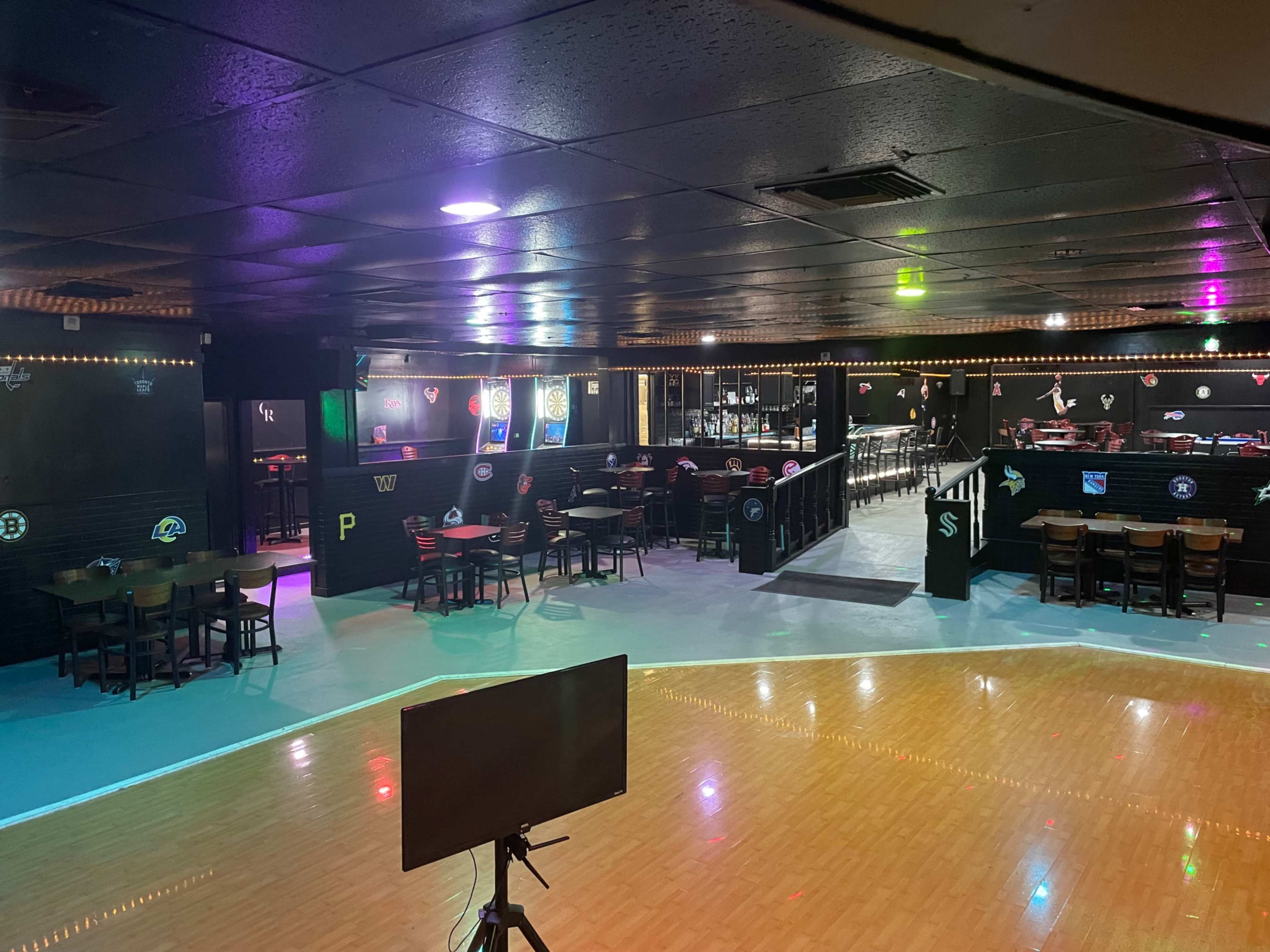 The image shows an empty indoor skating rink with tables and chairs scattered along the sides, dimly lit by colorful lights.