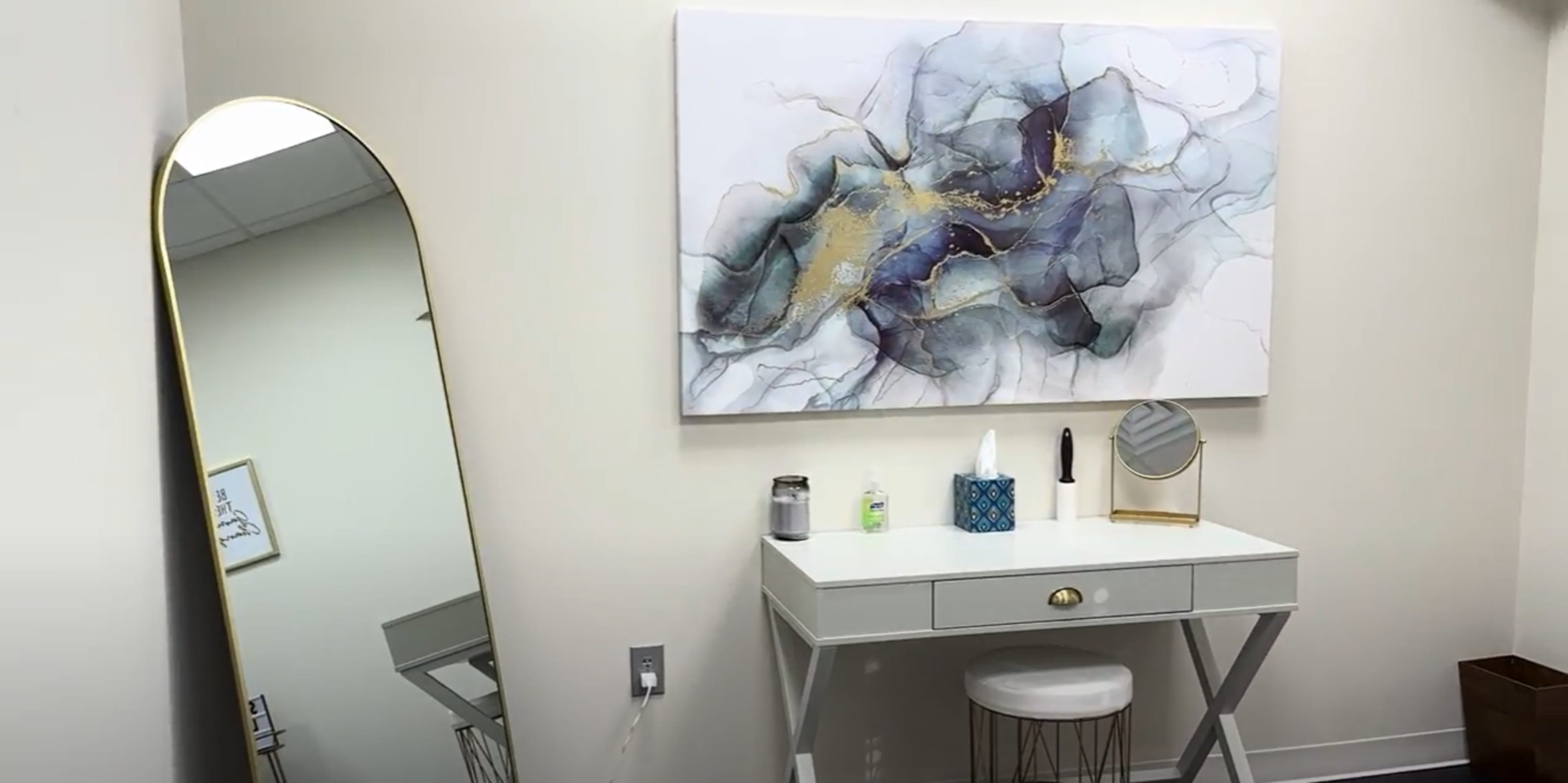 A stylish vanity area with a white desk, a round mirror, a decorative painting on the wall, and a tall, oval mirror nearby.