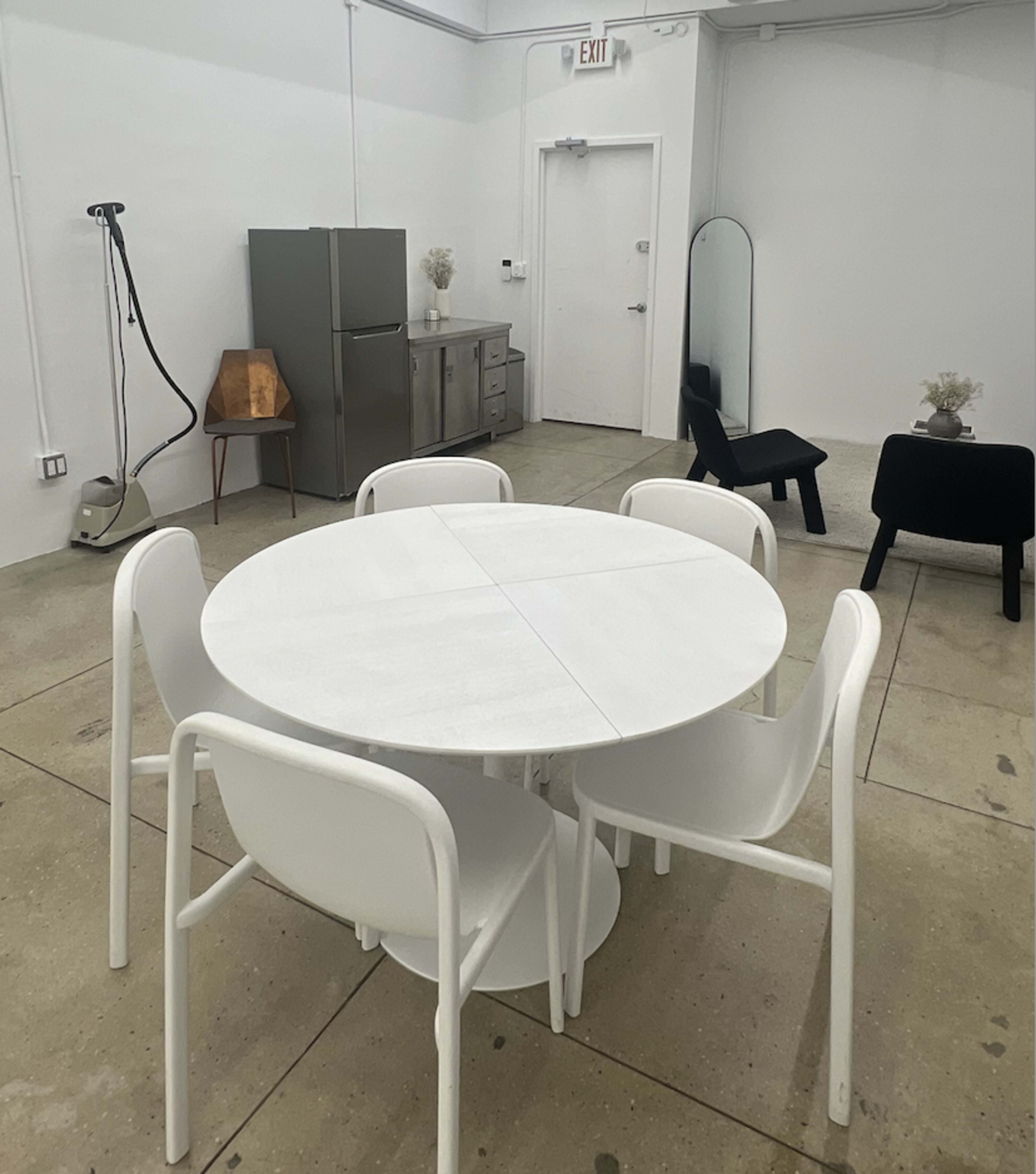 The image shows a minimalist kitchen area featuring a round white table surrounded by five white chairs, with a fridge, a small cabinet, and a mirror in the background.