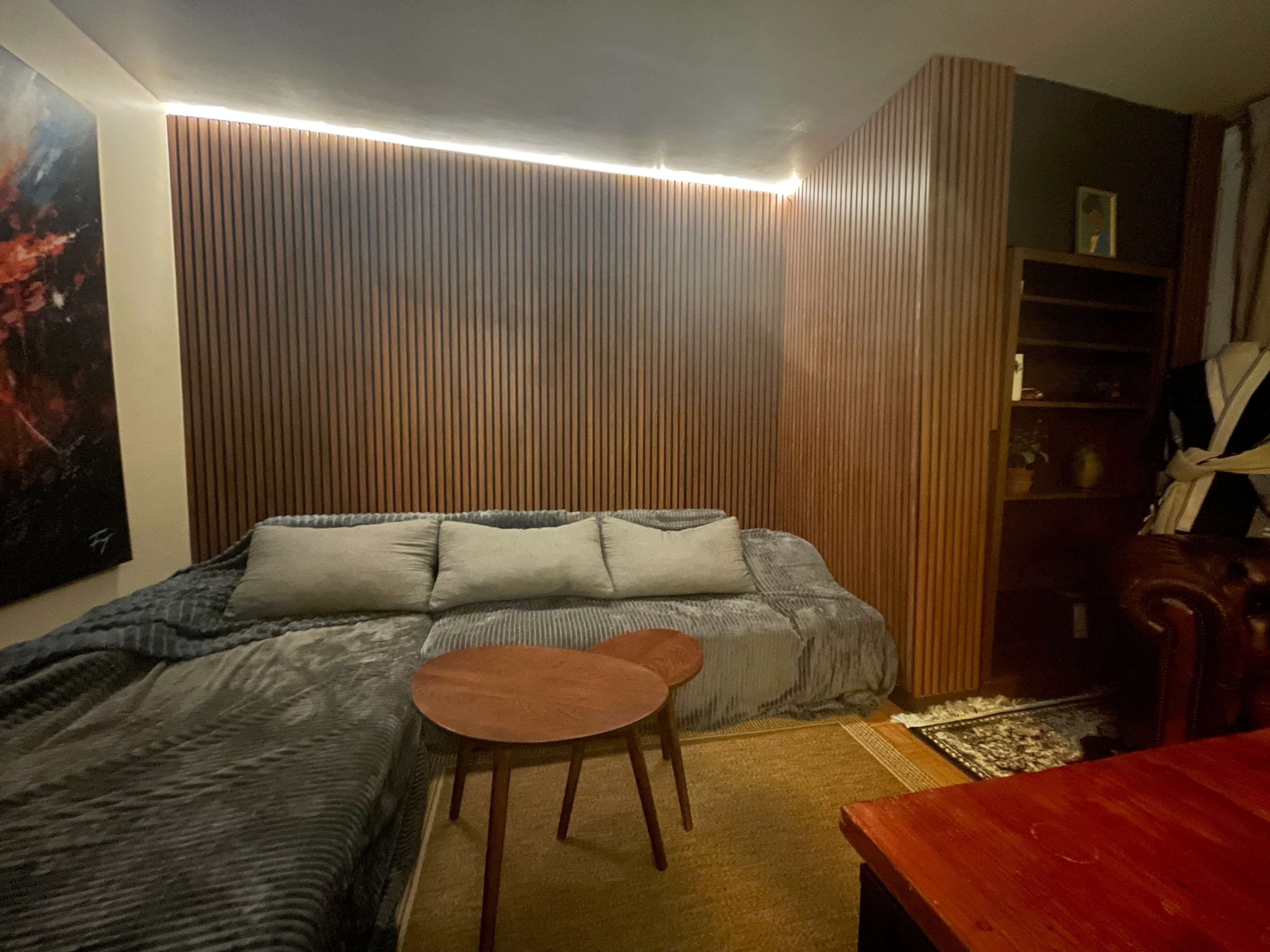 The image shows a cozy living room corner with a sectional sofa, a wooden coffee table, and a partially visible bookshelf against a wall with vertical wooden panels.