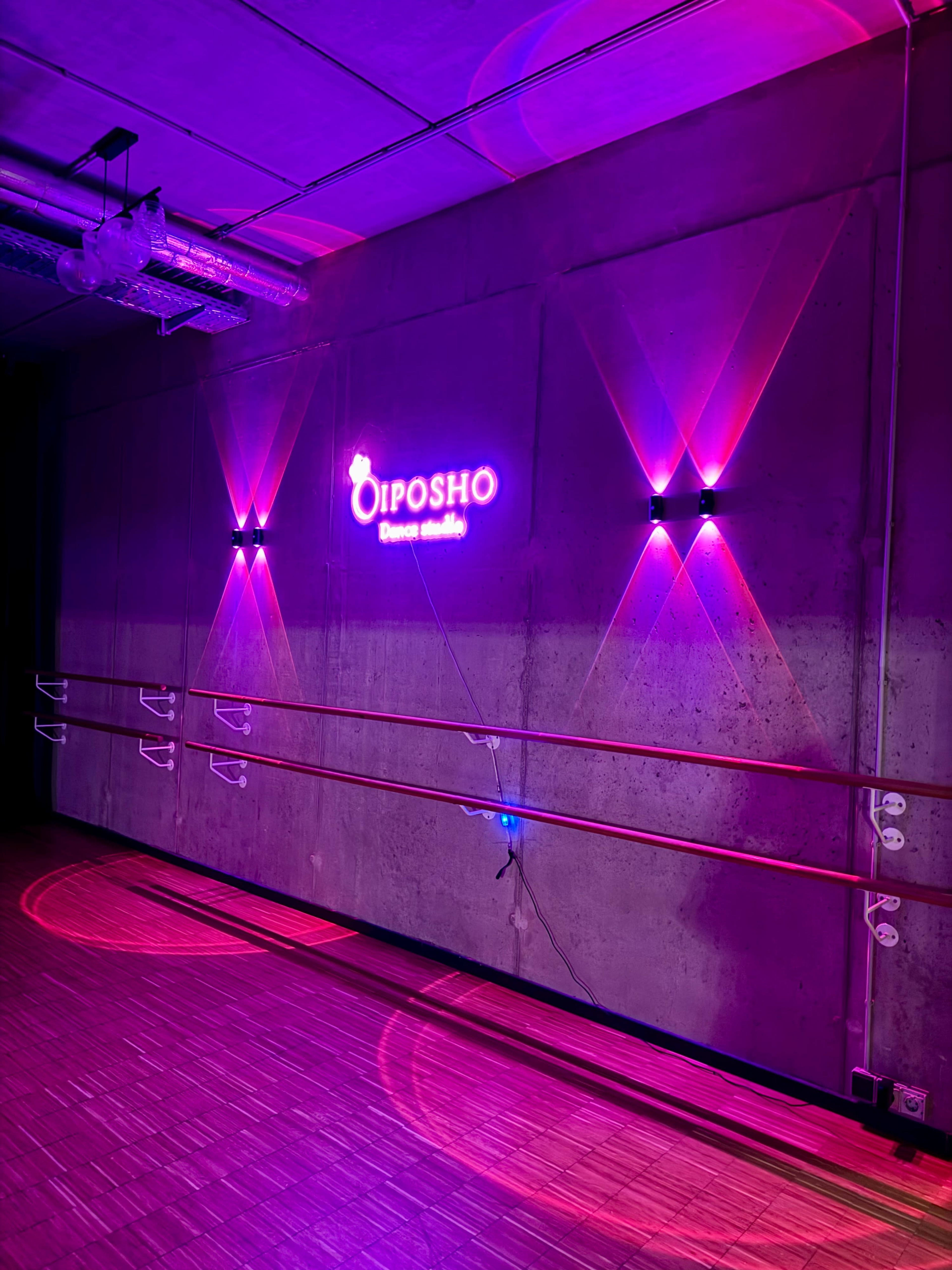 The interior of a dance studio features a concrete wall with the illuminated sign "OIPOSHO" and colored lights creating a vibrant ambiance.