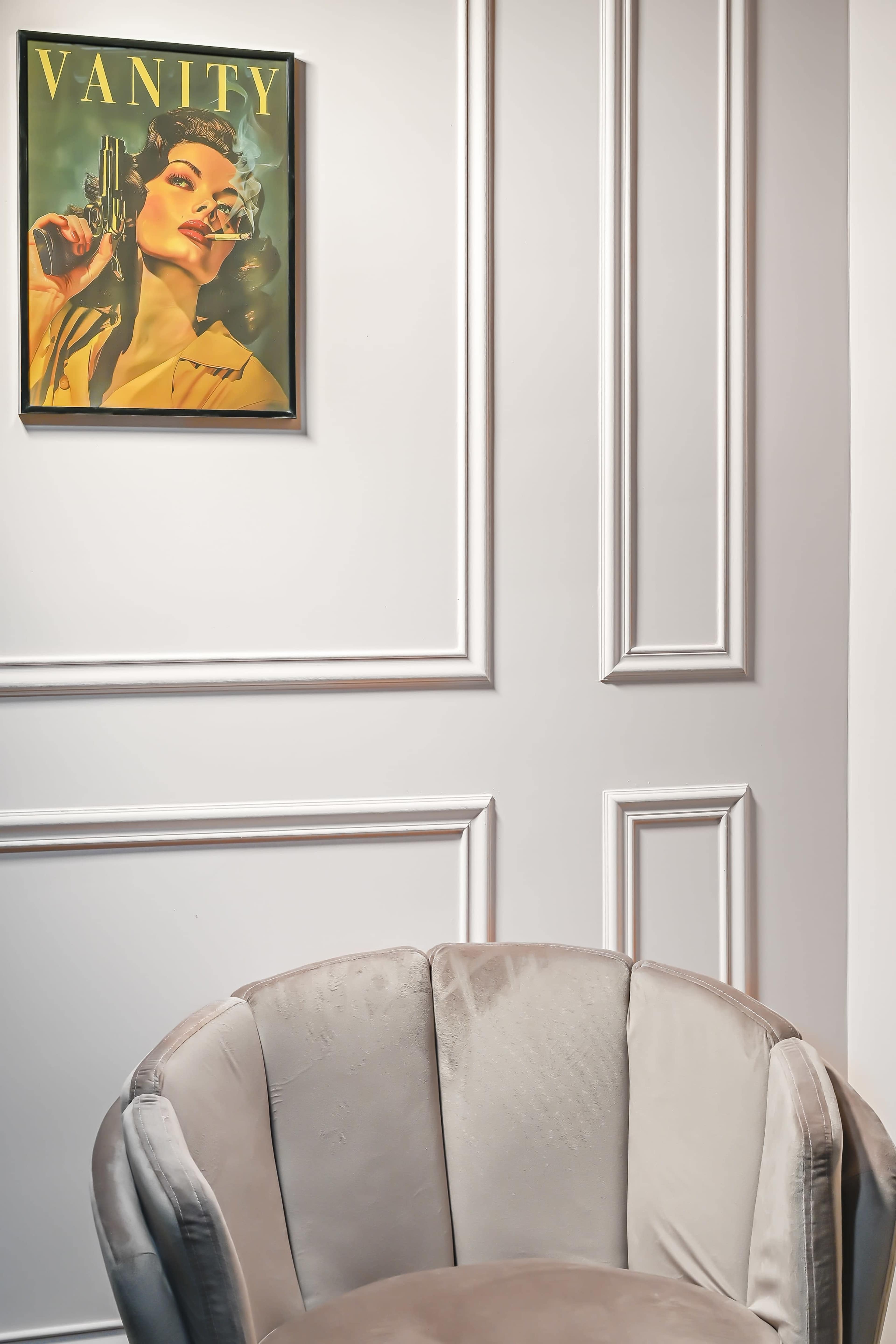 A vintage-inspired artwork titled "VANITY" hangs on a paneled wall above a plush, light-colored chair.