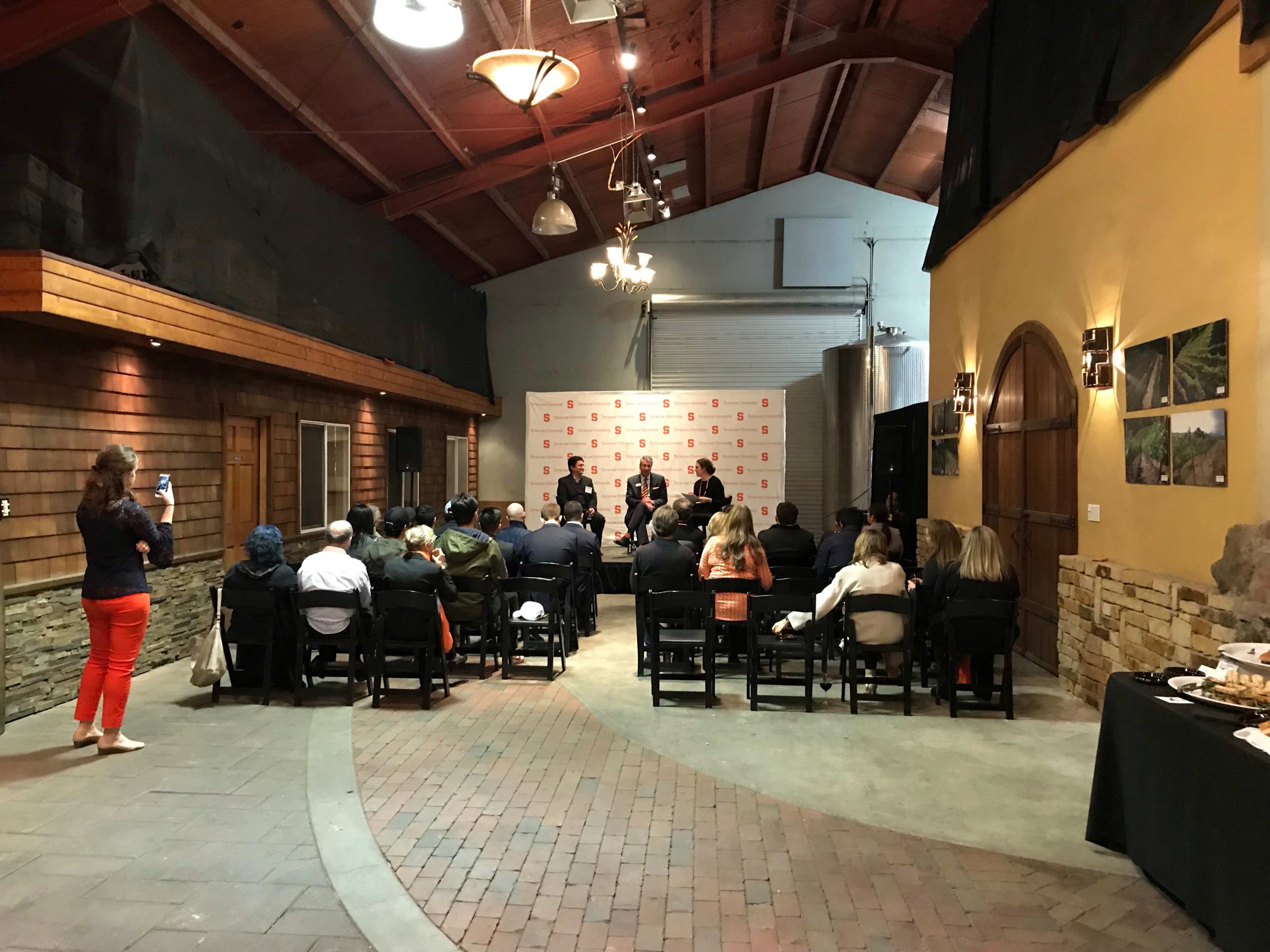 A group of people sits in chairs facing a panel at a presentation event indoors, with refreshments on a table to the side.