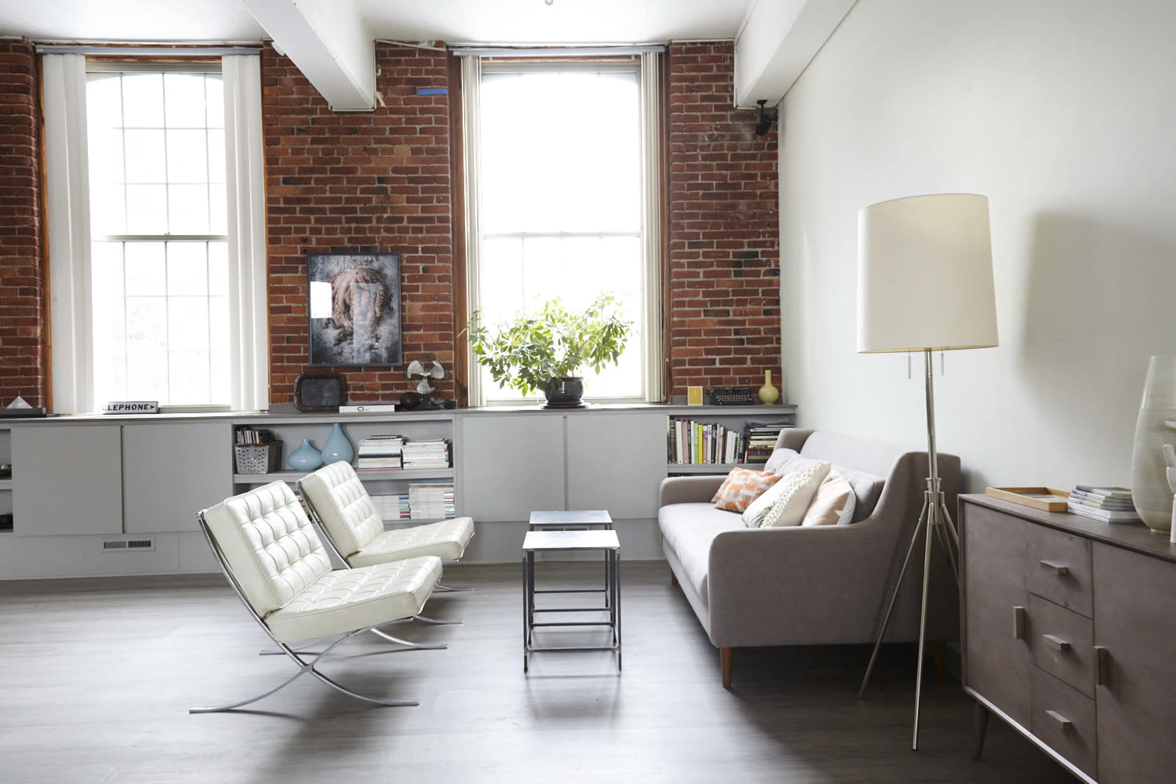A modern living room with a white chair and a gray sofa, featuring exposed brick walls and large windows, accompanied by a plant and shelves with books.