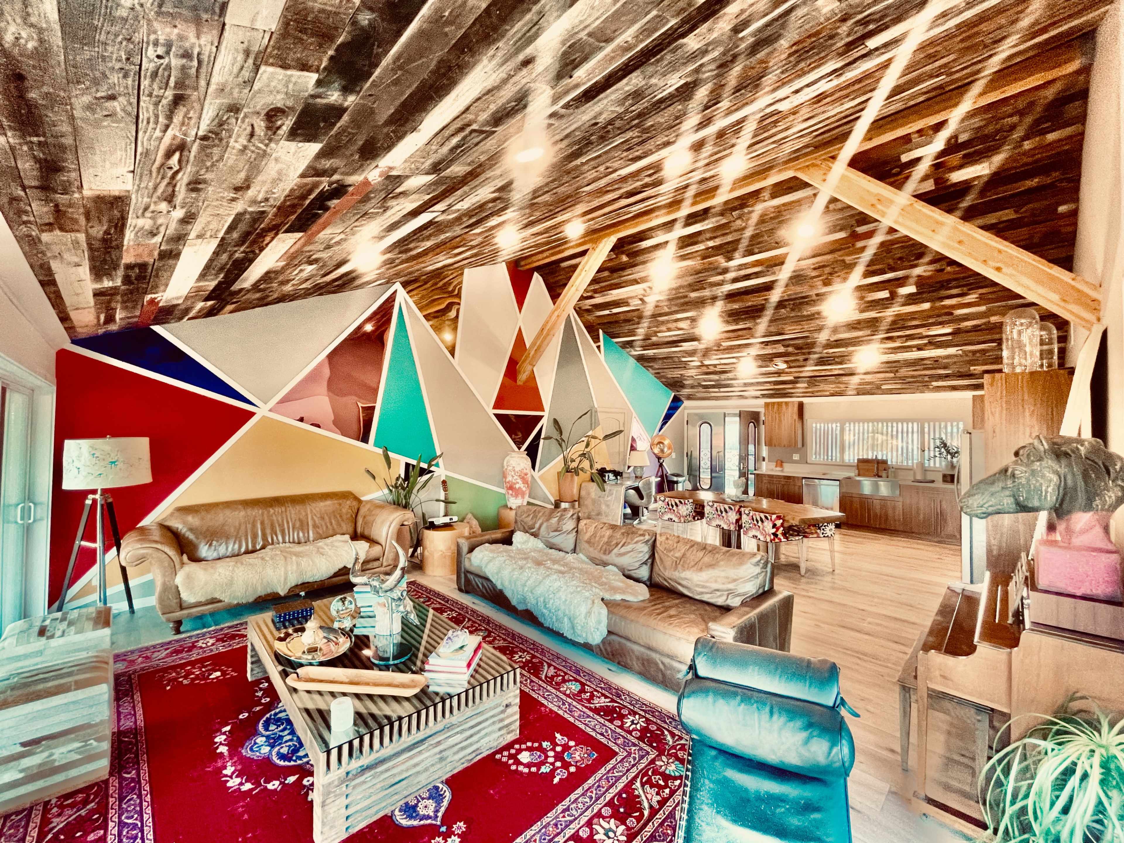 A spacious living room with a wooden ceiling, colorful geometric wall art, and various seating options arranged around a coffee table.