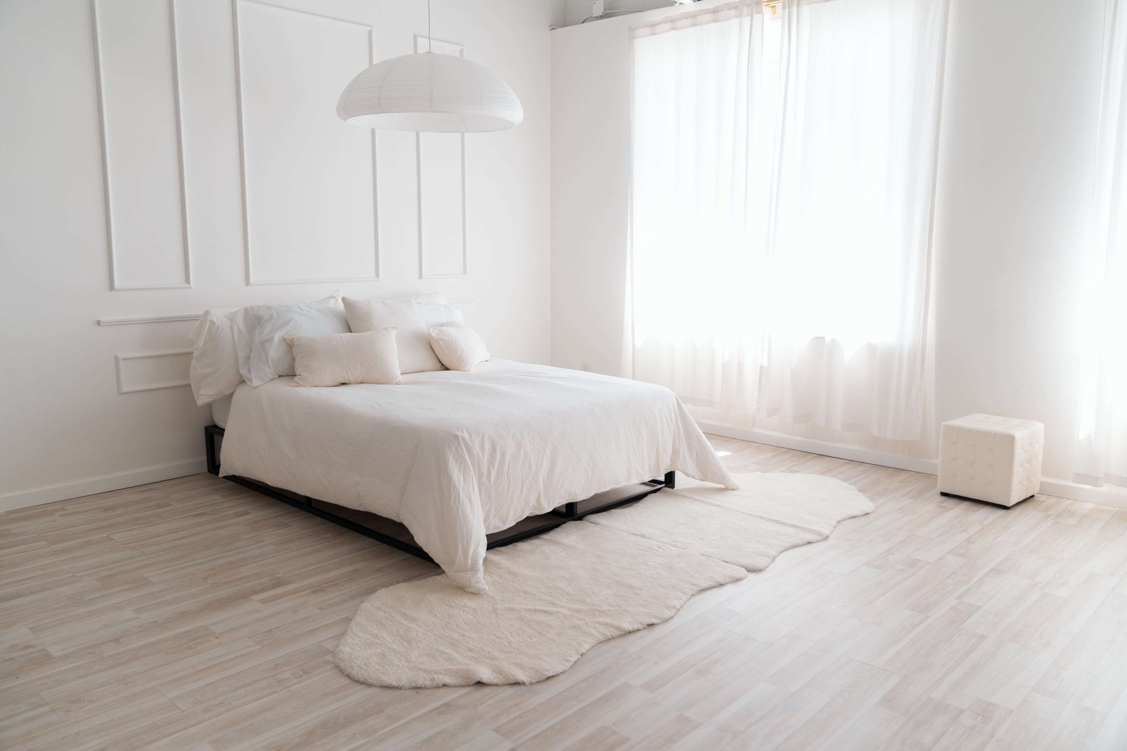 The image shows a minimalist bedroom featuring a white bed with cushions, a round pendant light, and large windows allowing natural light to enter.