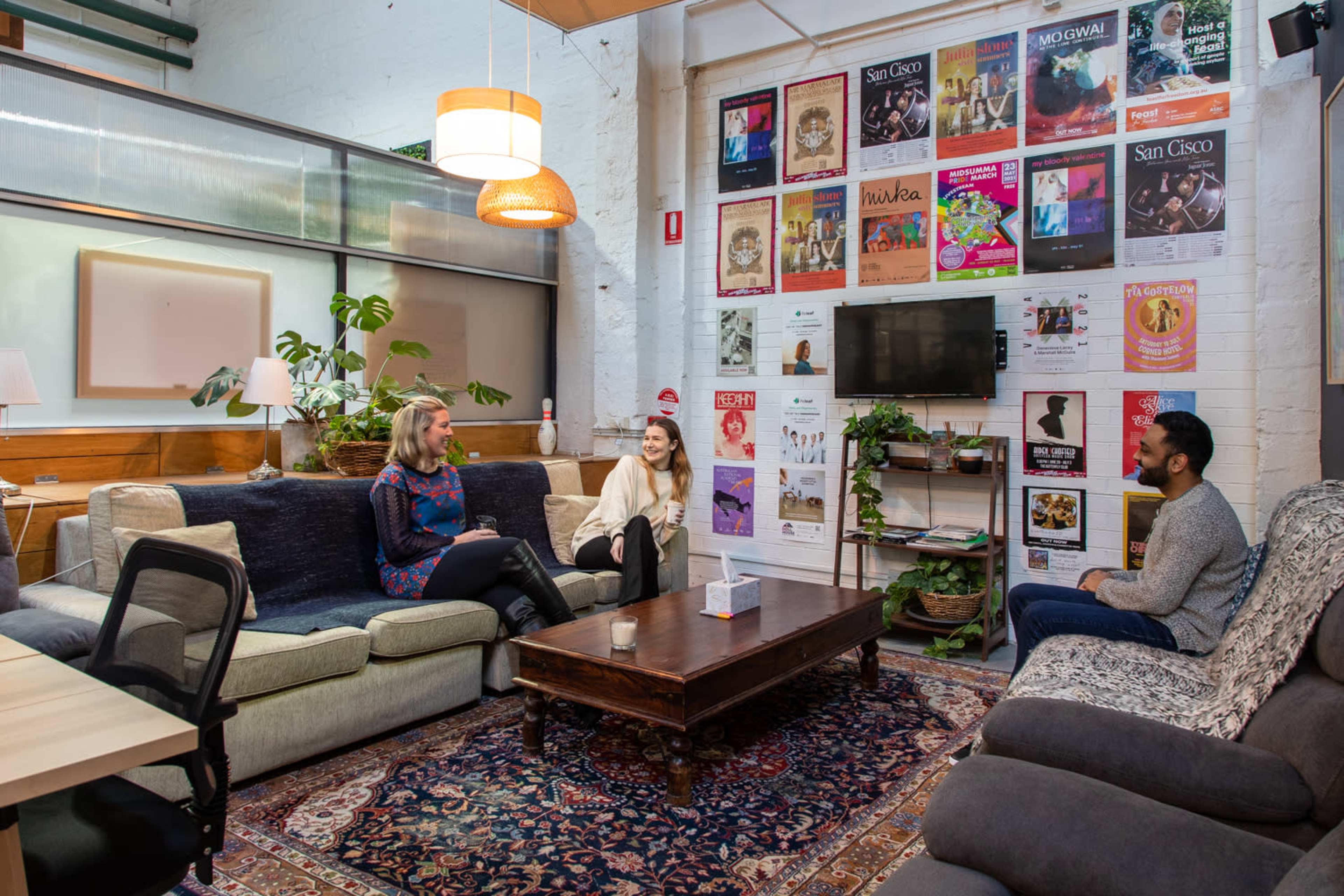 Three people are seated in a cozy lounge area with a large rug, surrounded by potted plants and a wall adorned with various event posters.