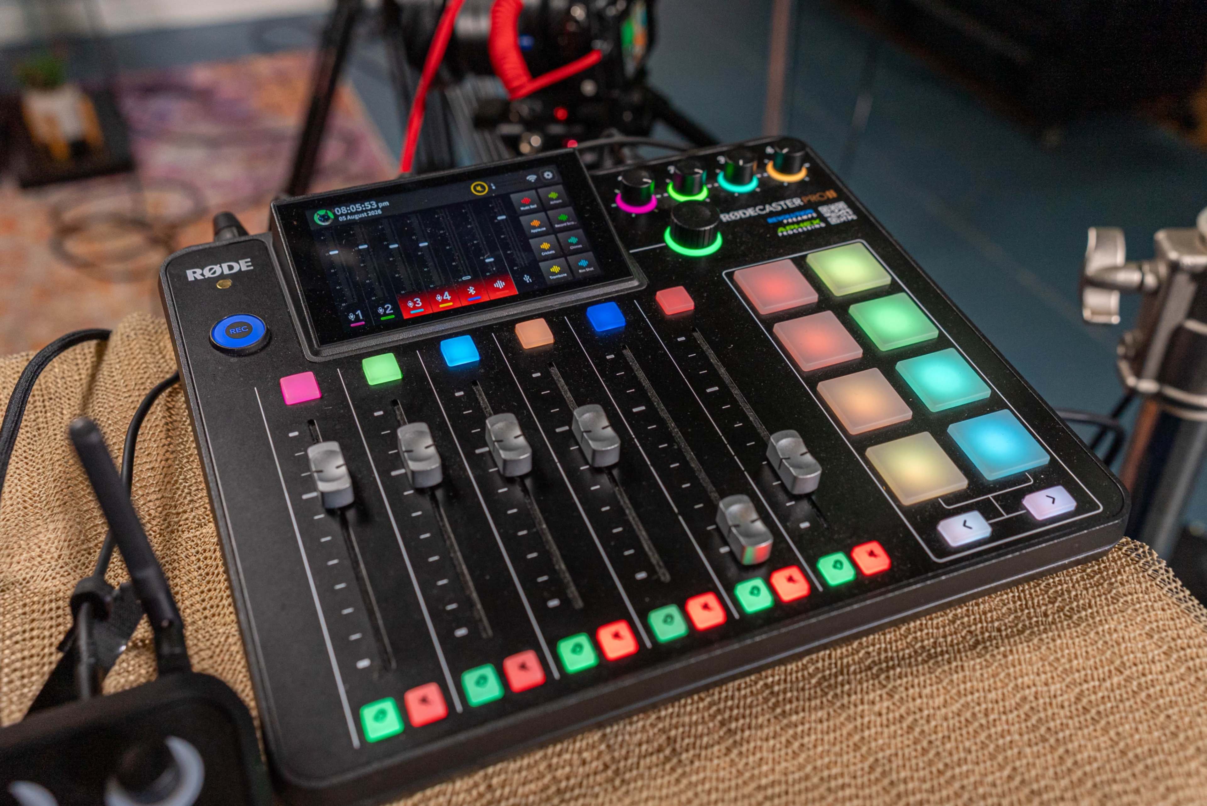 The image shows a Rode audio mixer with multiple sliders, buttons, and a touchscreen display, set on a textured surface.