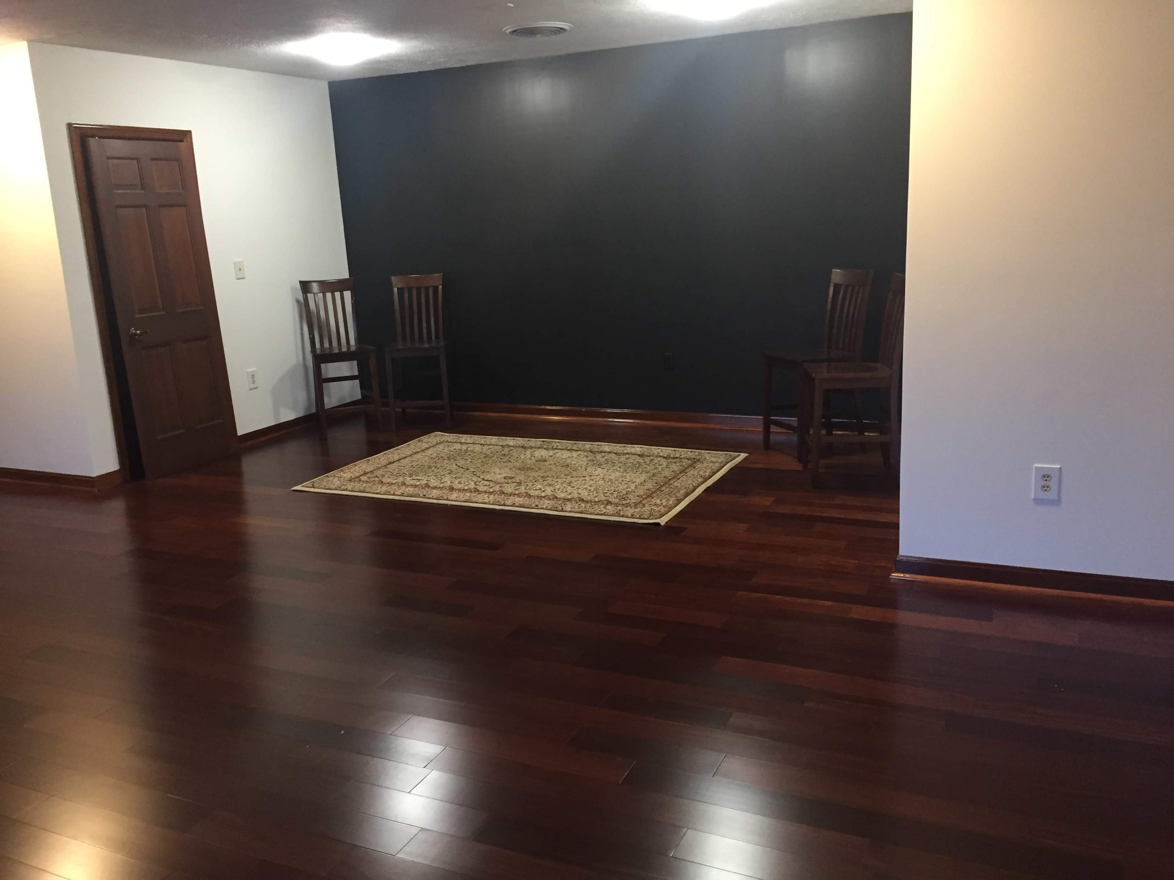 The image shows a room with dark wooden flooring, a patterned rug in the center, two wooden chairs on one side, and a dark feature wall.