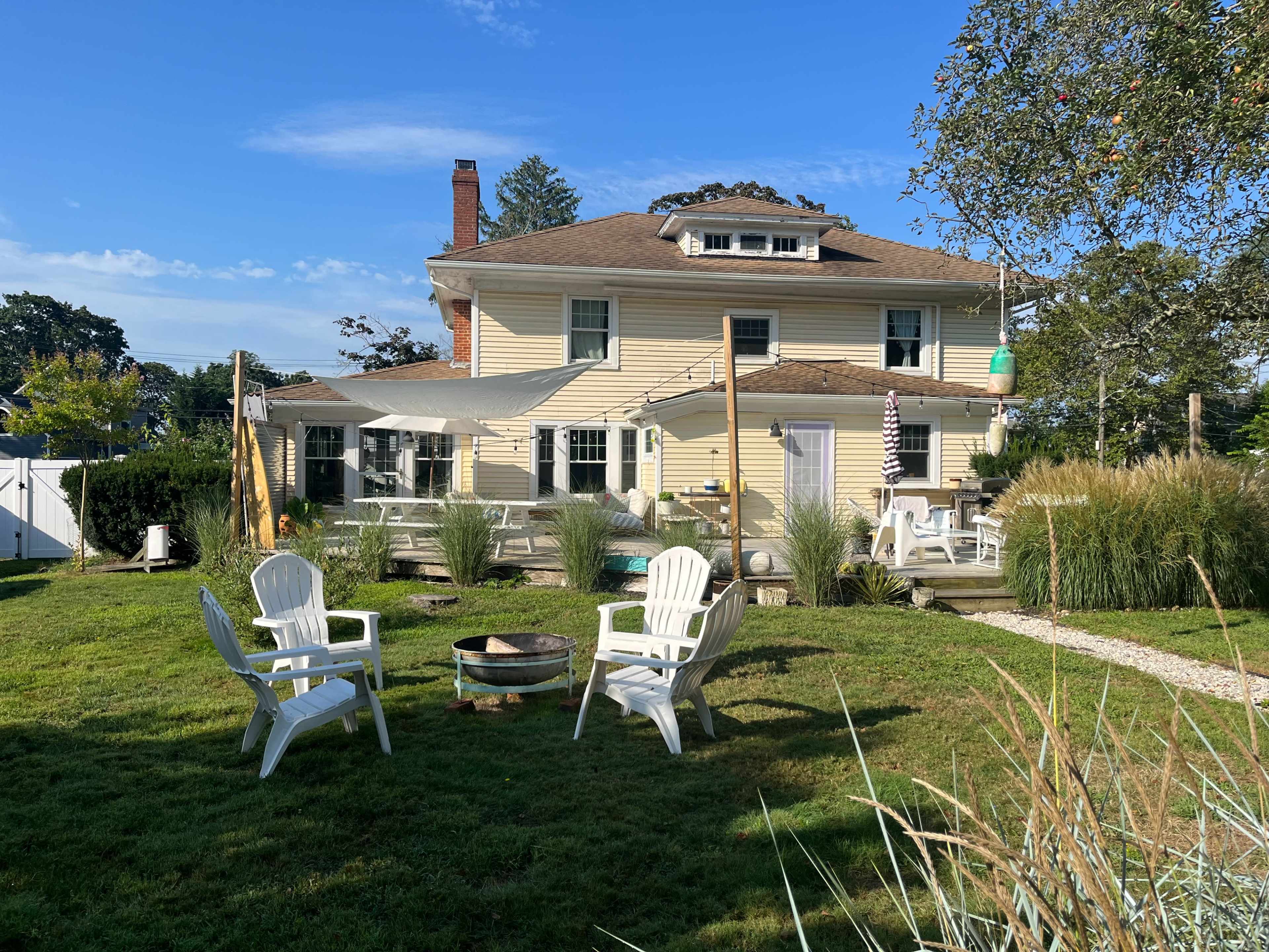 A large yellow house with a broad porch and a fire pit surrounded by white chairs sits in a grassy yard under a clear blue sky.