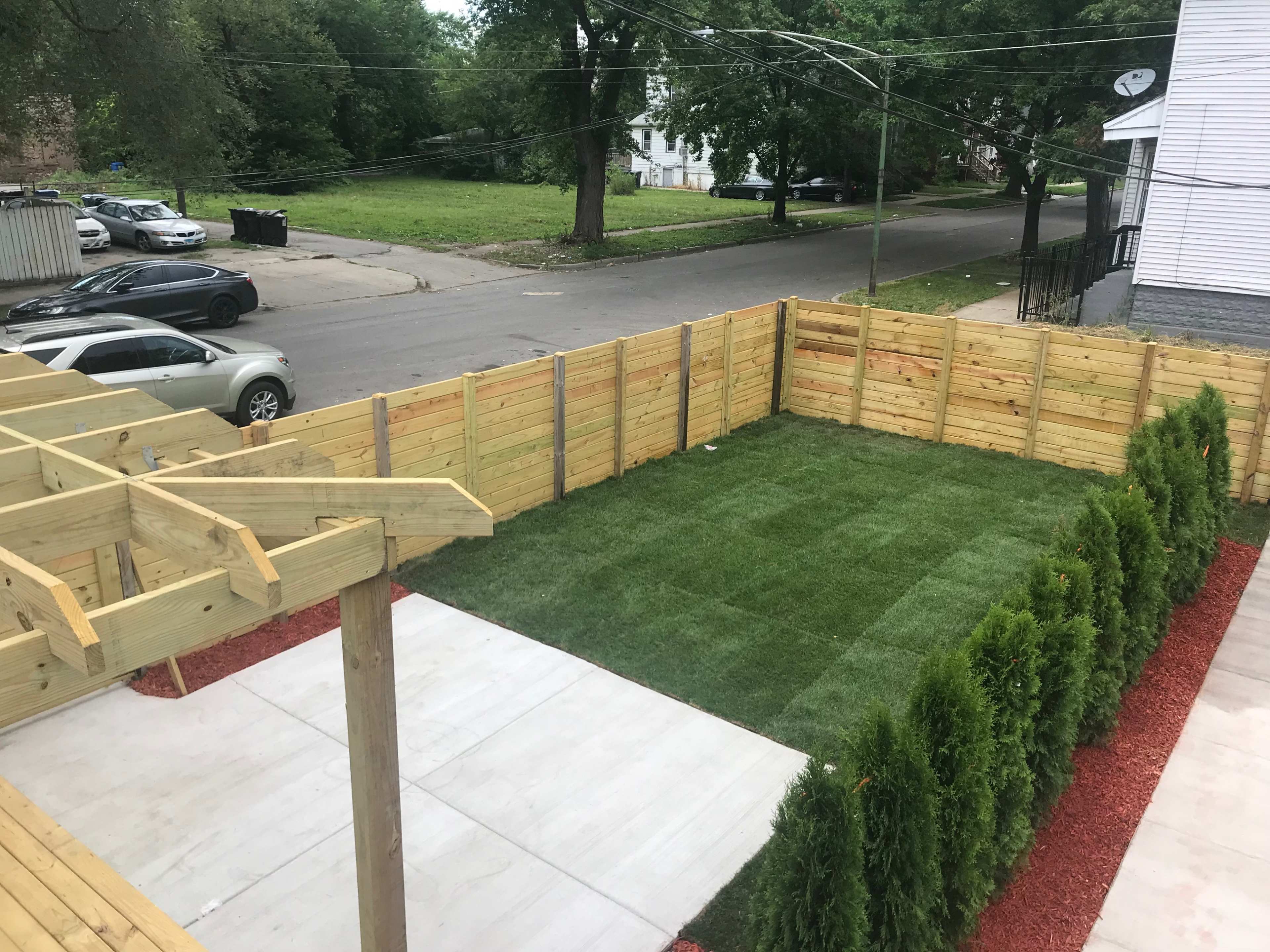 A fenced backyard features manicured grass and a concrete area, bordered by small trees and adjacent to a street with parked cars.