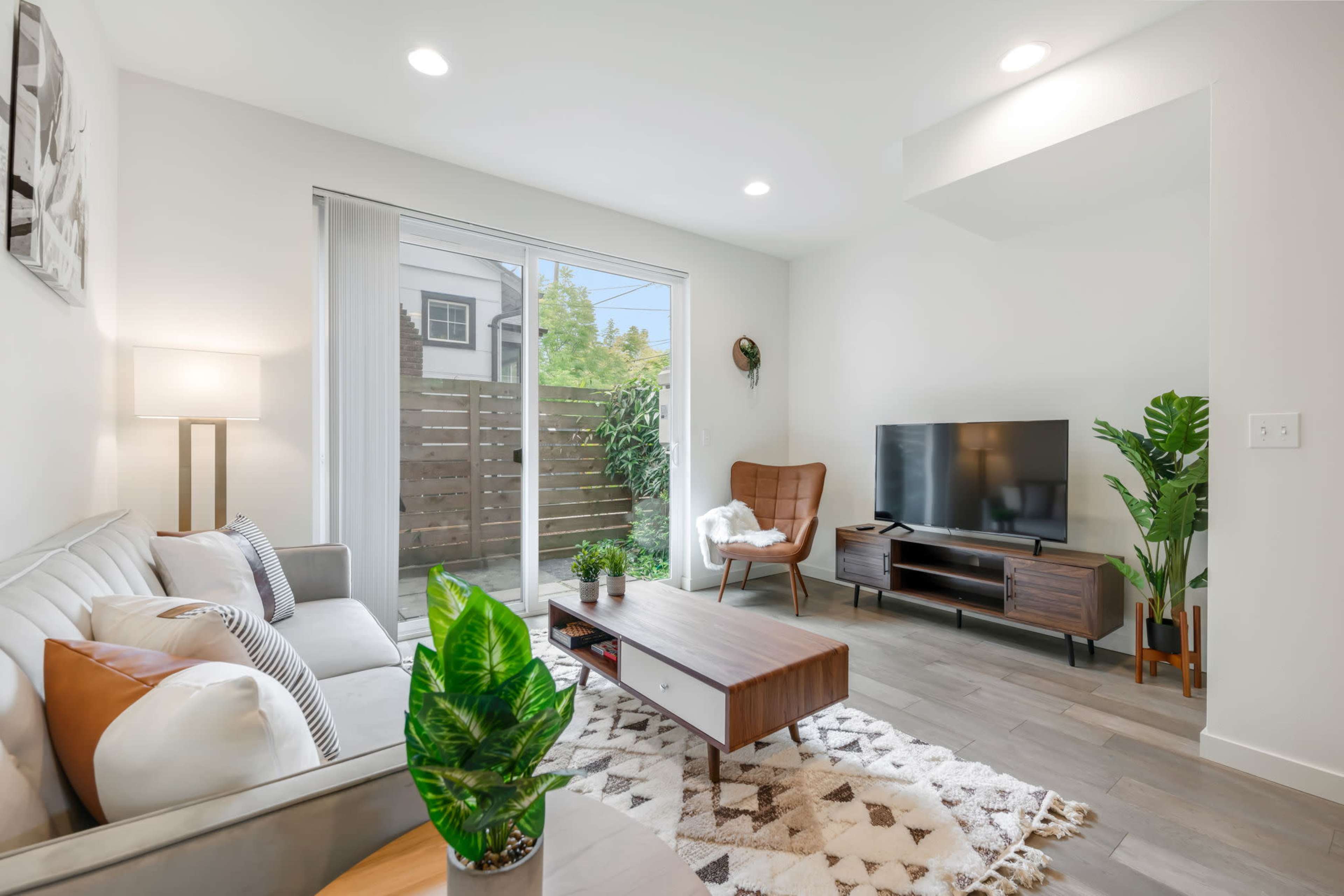 A modern living room featuring a gray sofa, a wooden coffee table, a television stand with a TV, and a large window overlooking a small outdoor space.