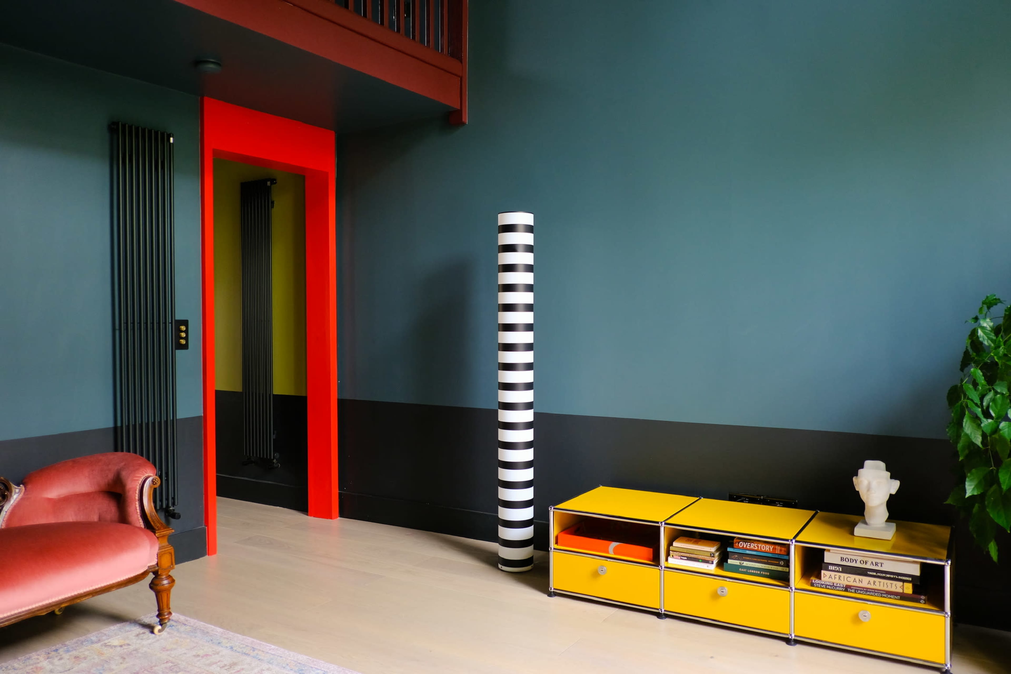 The room features a pink upholstered chair, a black and white striped column, a yellow storage unit with books, and a bold red doorway against dark green walls.