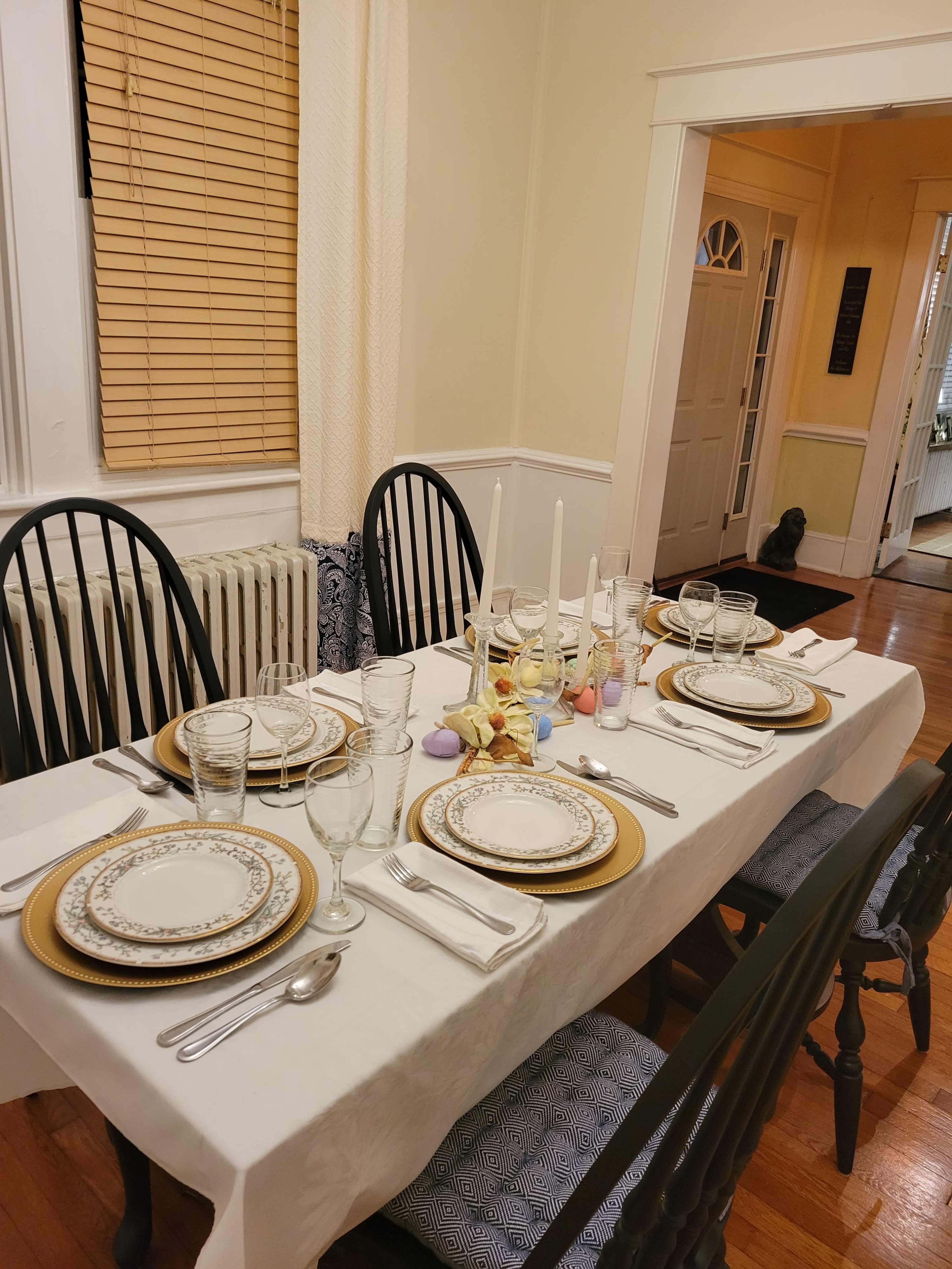 A dining table is set with plates, glassware, and cutlery, arranged neatly for a meal in a well-lit room with a doorway leading to another area.