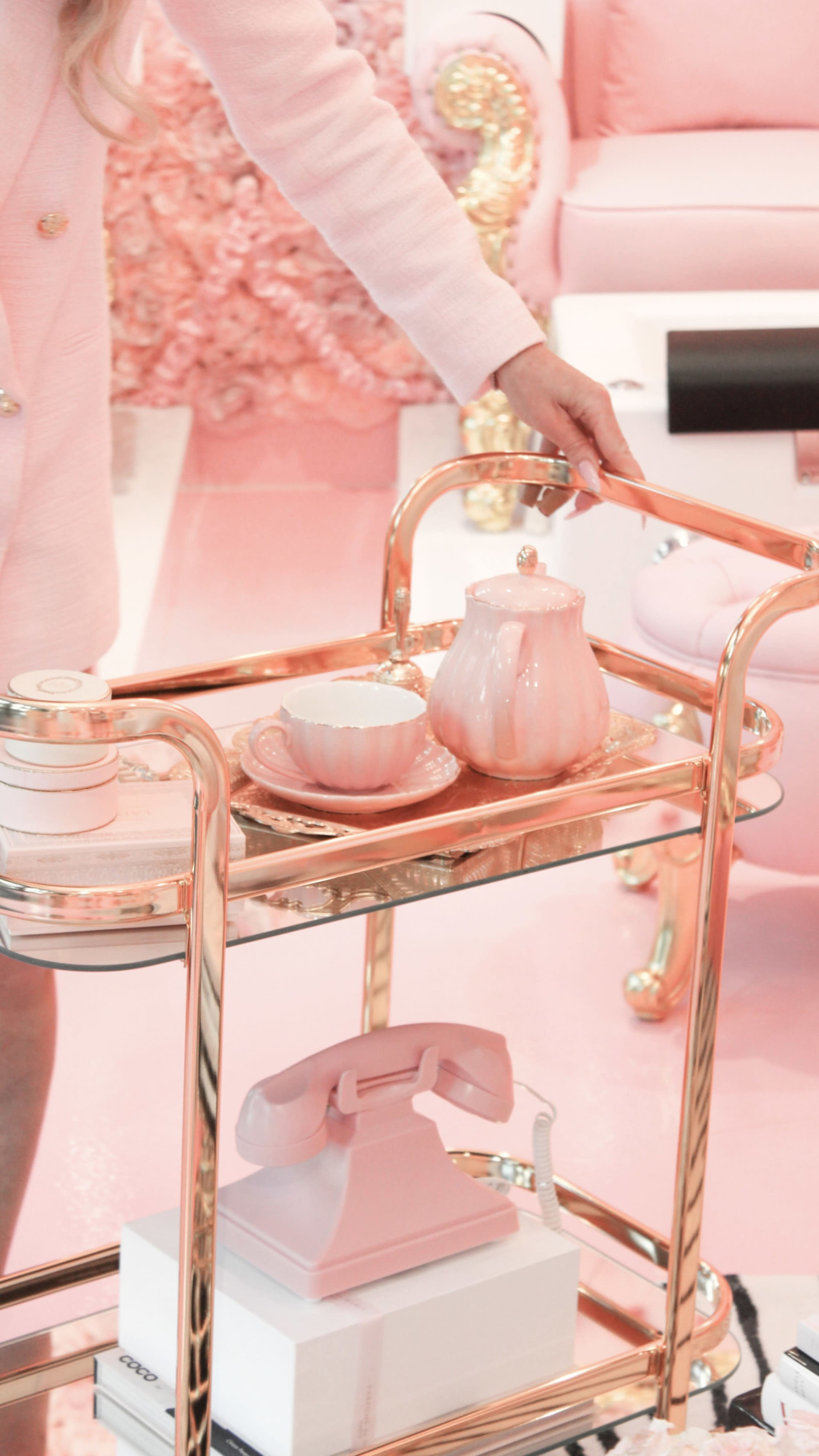 A person holds a gold and glass serving cart adorned with pink tea set items on a pink floor.