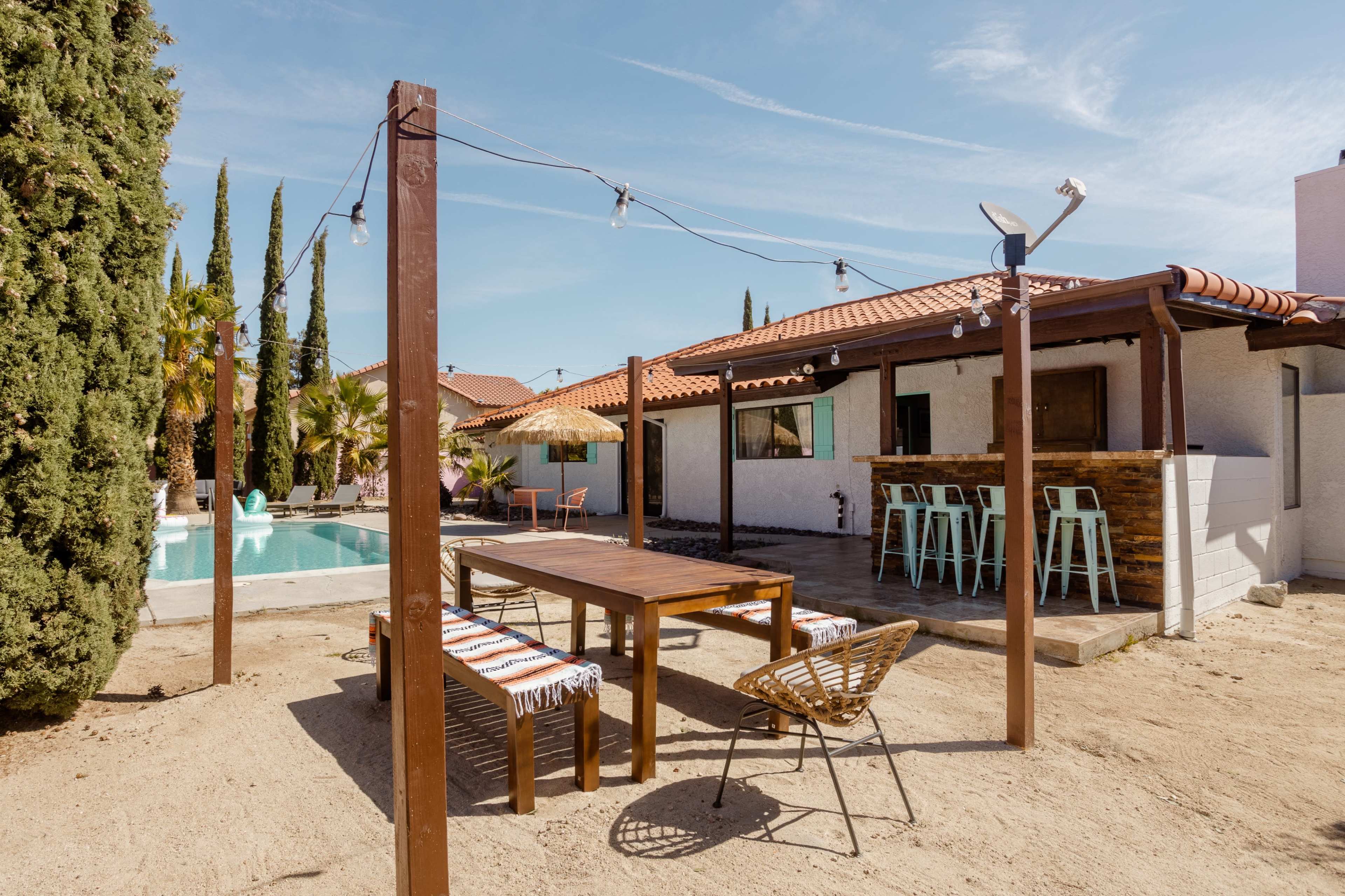 A wooden patio with a table and seating area overlooks a swimming pool, surrounded by tall cypress trees and a house in the background.