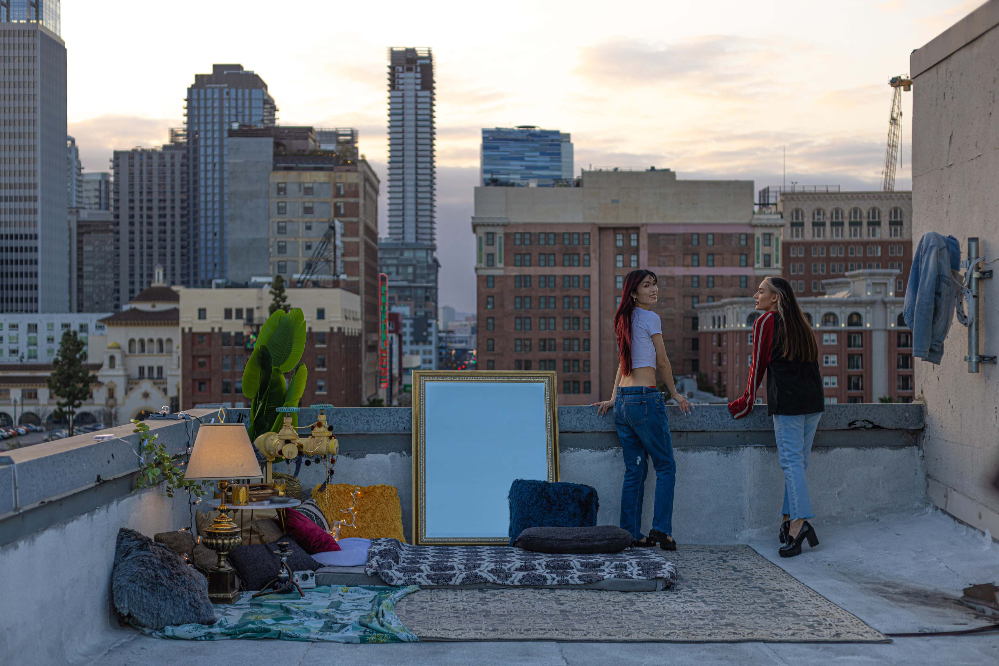 Two women stand on a rooftop decorated with rugs, a large mirror, a lamp, and plants, with a city skyline visible in the background during twilight.