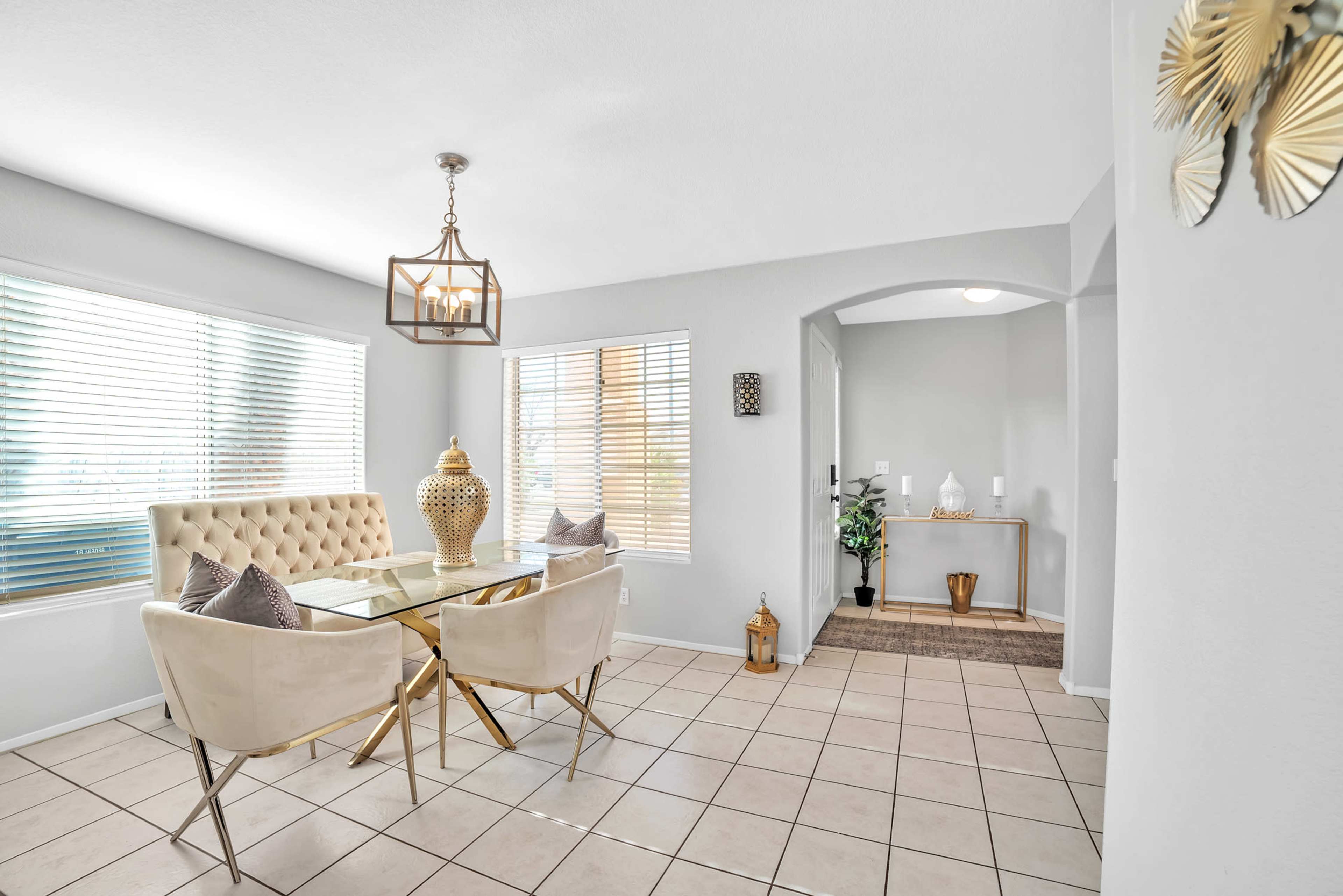 A dining area with a glass table surrounded by beige chairs, large windows with blinds, and a decorative entrance visible in the background.
