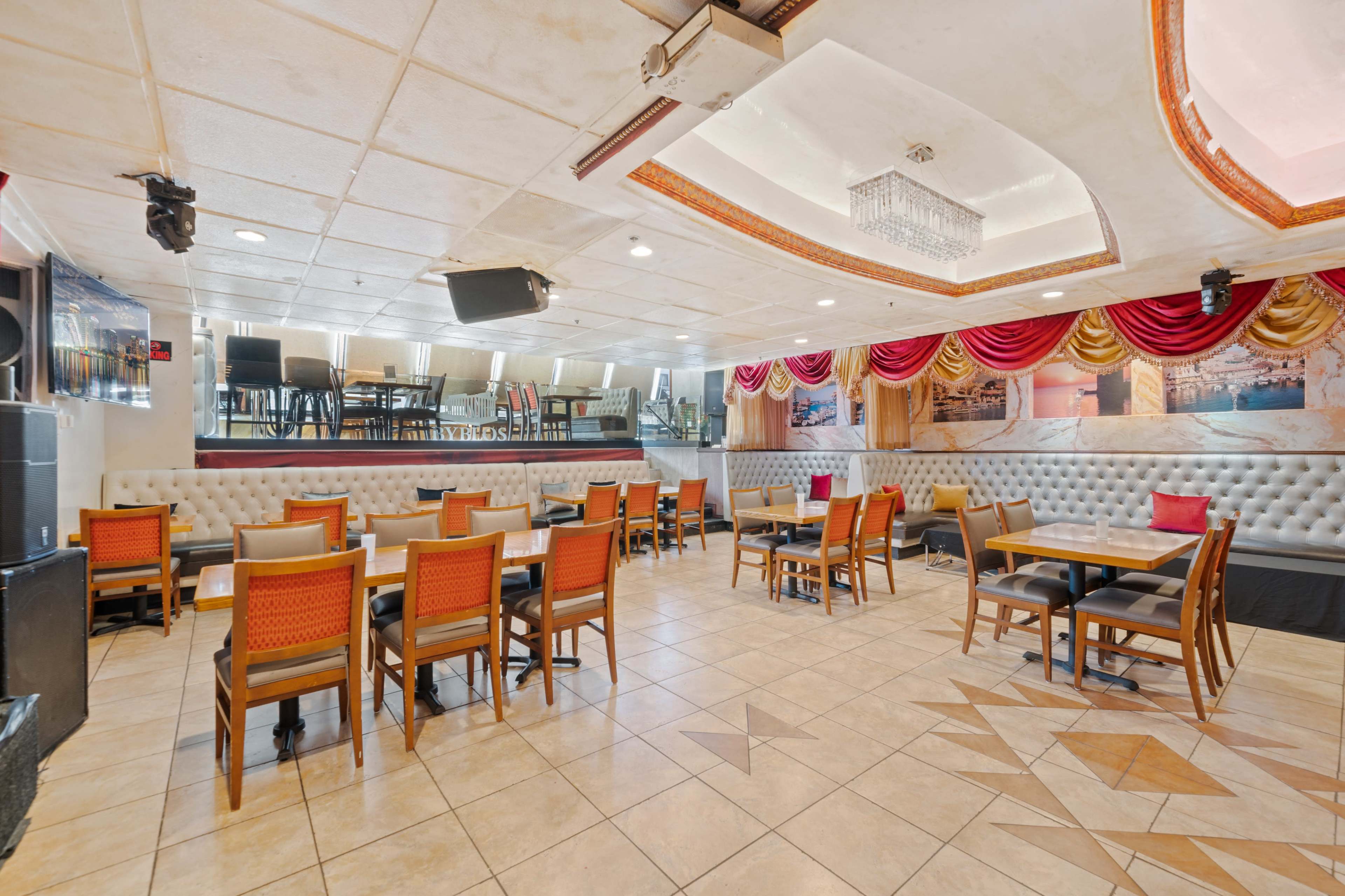 An empty restaurant interior with tables and chairs arranged on a tiled floor, adorned with red drapery and decorative wall panels.