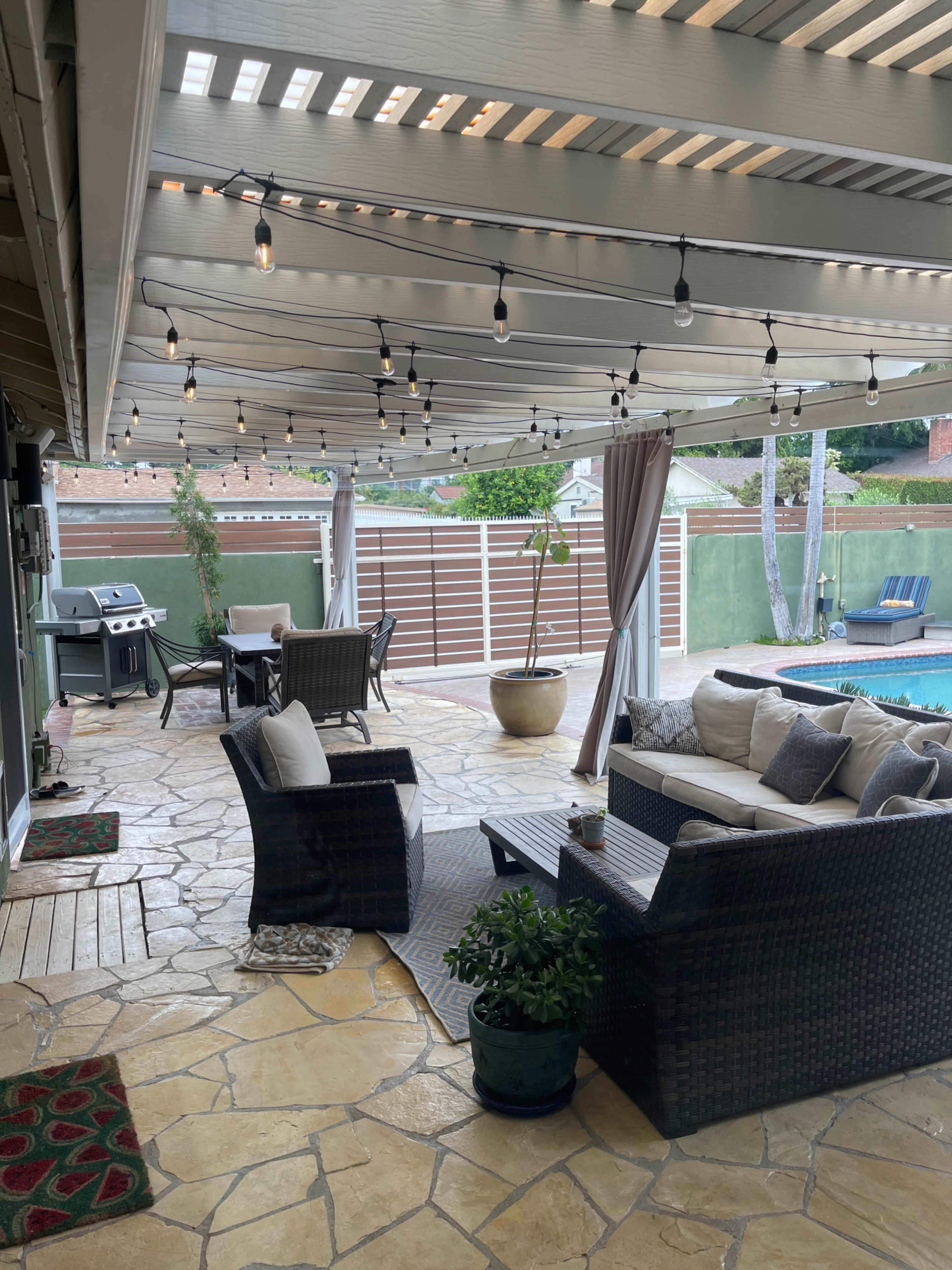 A patio area with a stone floor, outdoor furniture, a grill, and decorative string lights overhead, adjacent to a swimming pool.