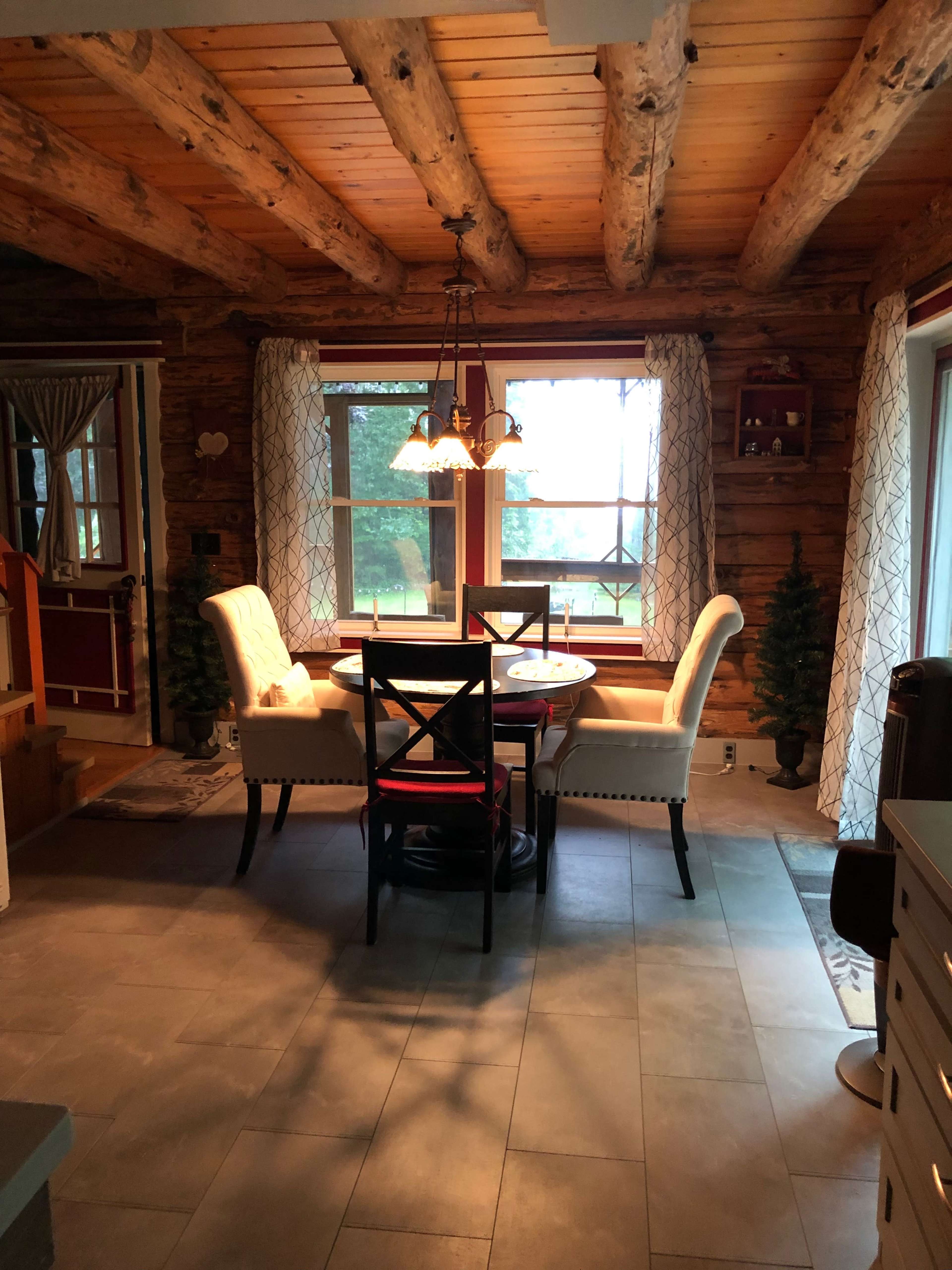 A rustic log cabin interior features a dining table with chairs near large windows, providing natural light and views of the outdoors.