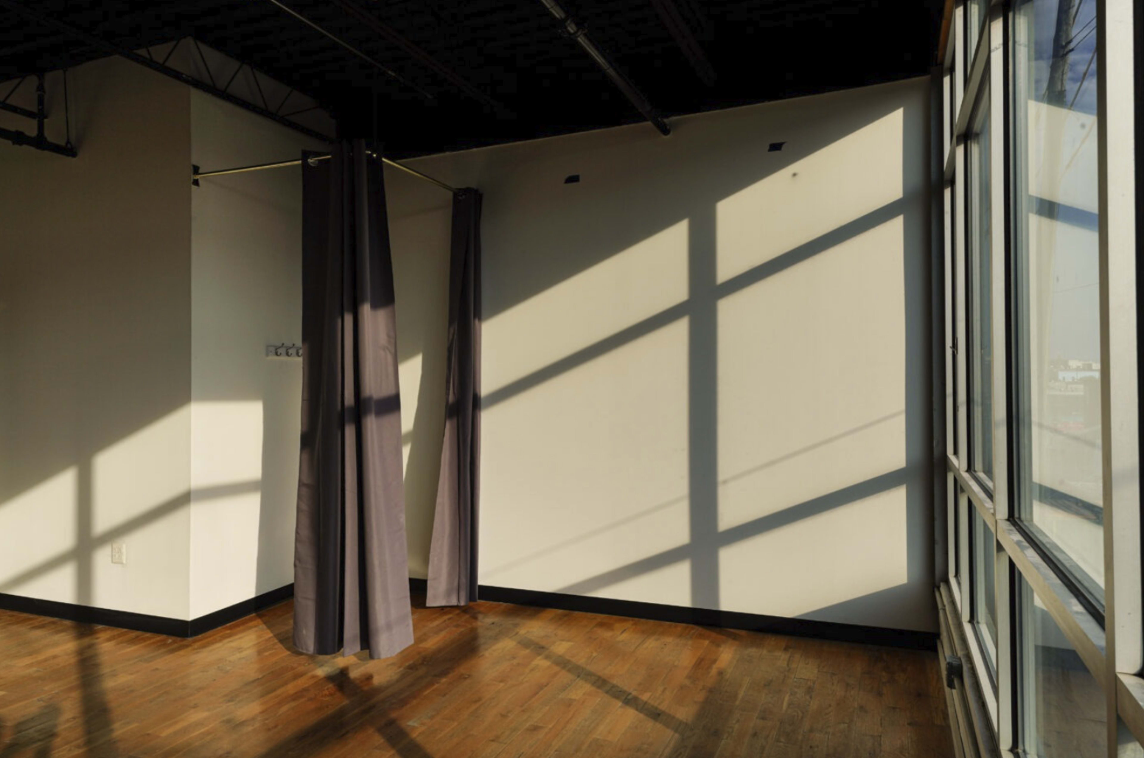 A sunlit interior of a room features a wooden floor, a large window, and gray curtains hanging near the wall.
