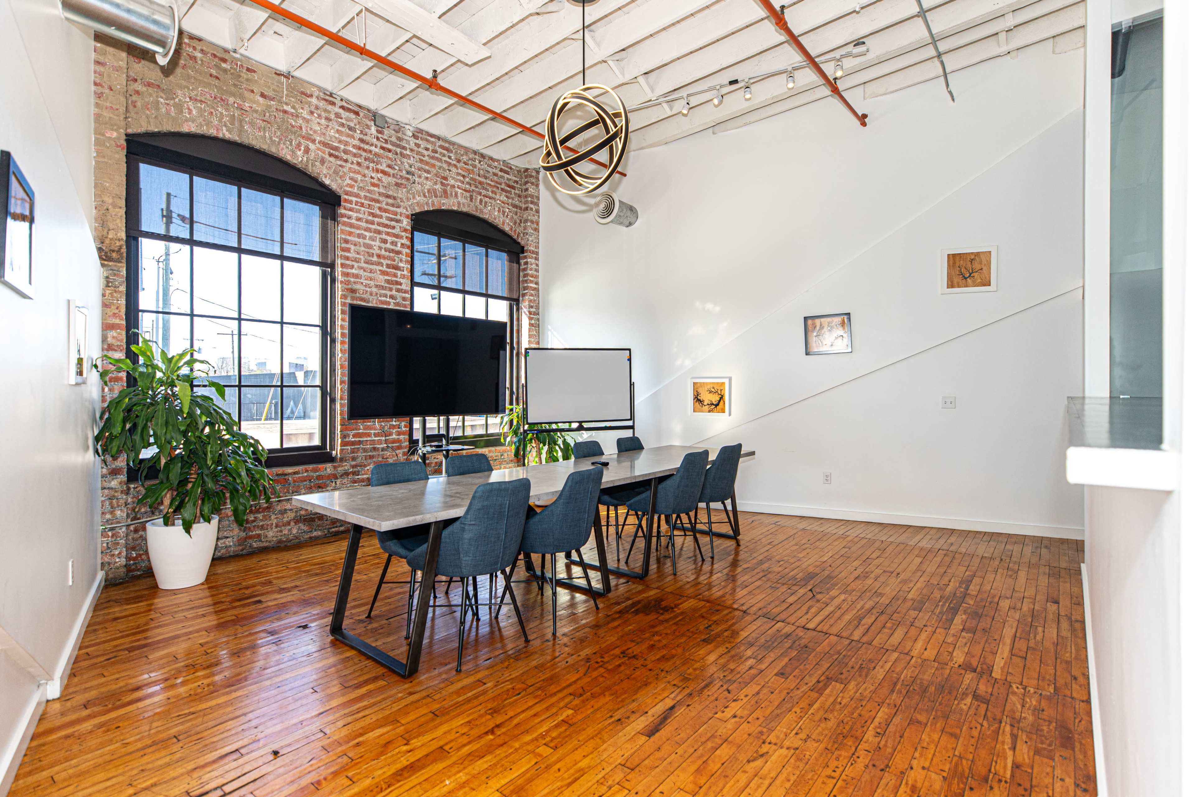 A spacious conference room features a long table surrounded by chairs under industrial-style windows, with exposed brick walls and wooden floors.