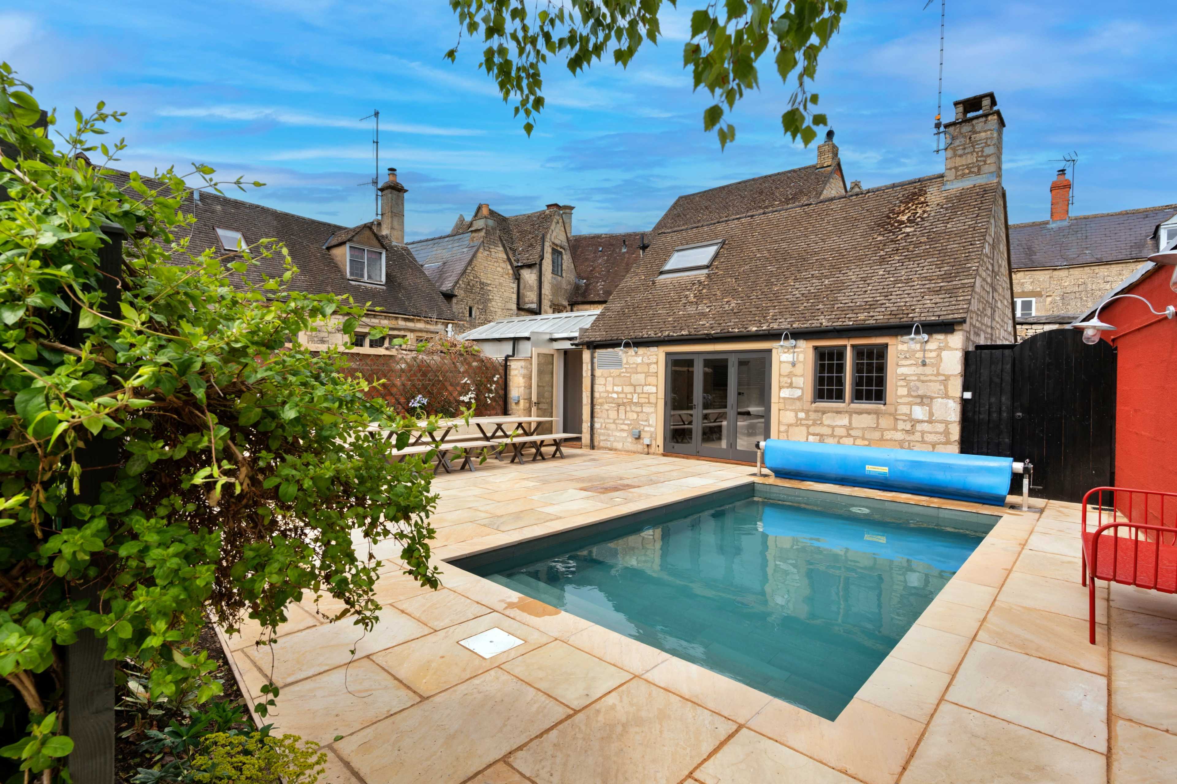 A small rectangular swimming pool surrounded by stone tiles and a seating area adjacent to historical-style buildings.