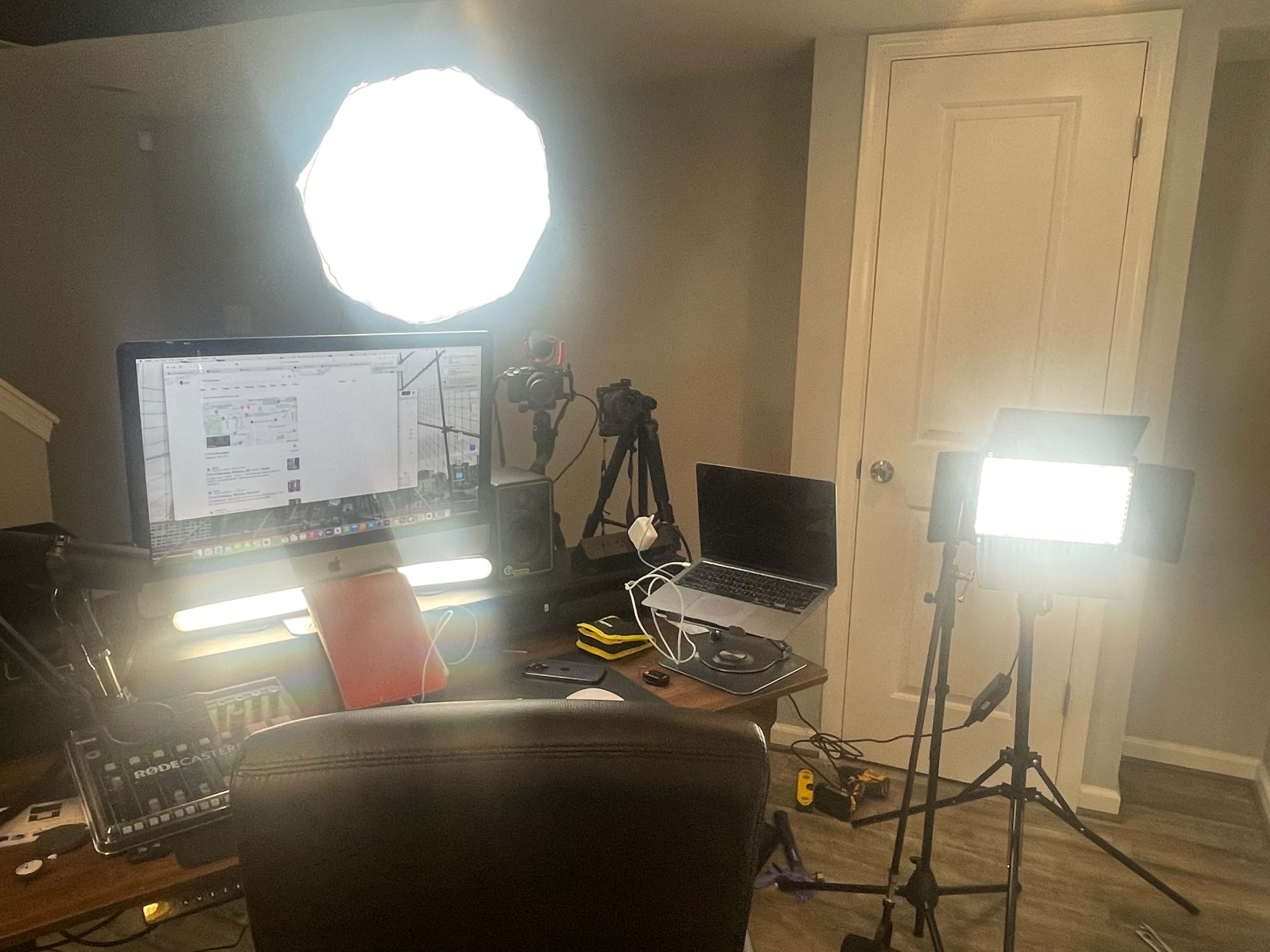 A cluttered workspace features multiple cameras, a computer monitor displaying a webpage, and two bright lights set up for video production.