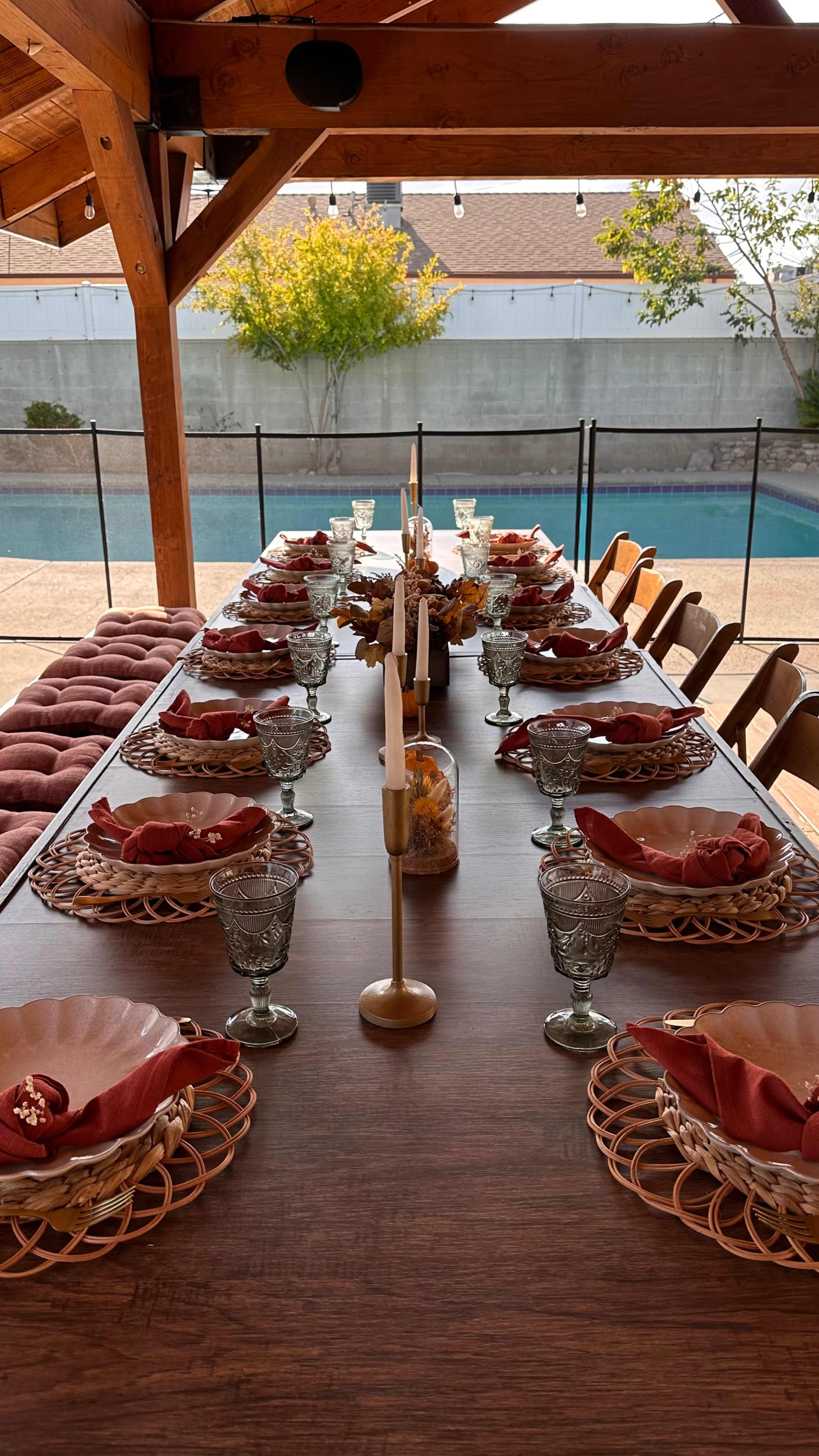 A long dining table is set outdoors with decorative plates, glasses, and centerpieces, overlooking a swimming pool.