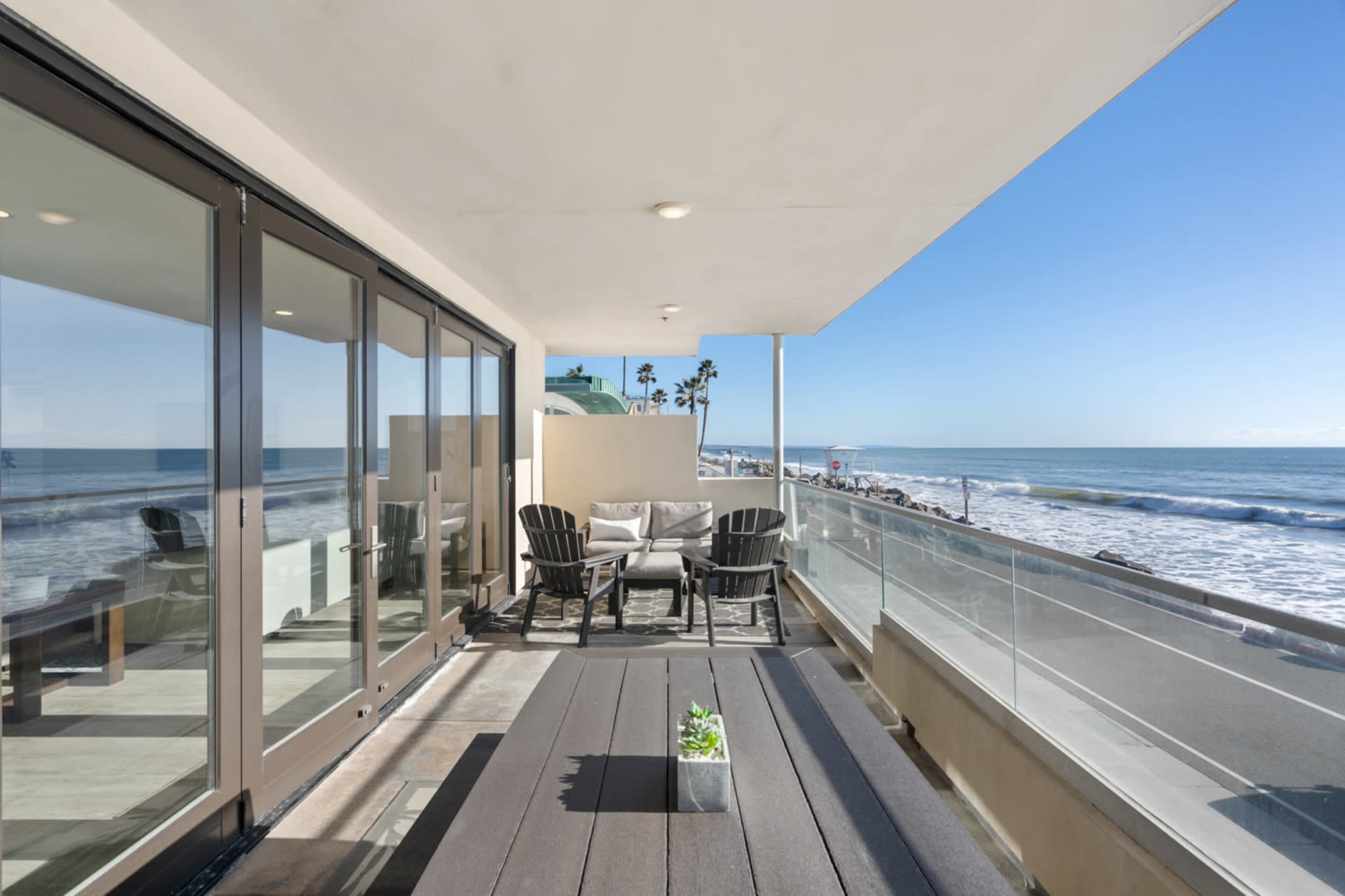 A balcony overlooking the ocean features a seating area with chairs and a table, surrounded by a glass railing with views of the beach and palm trees.