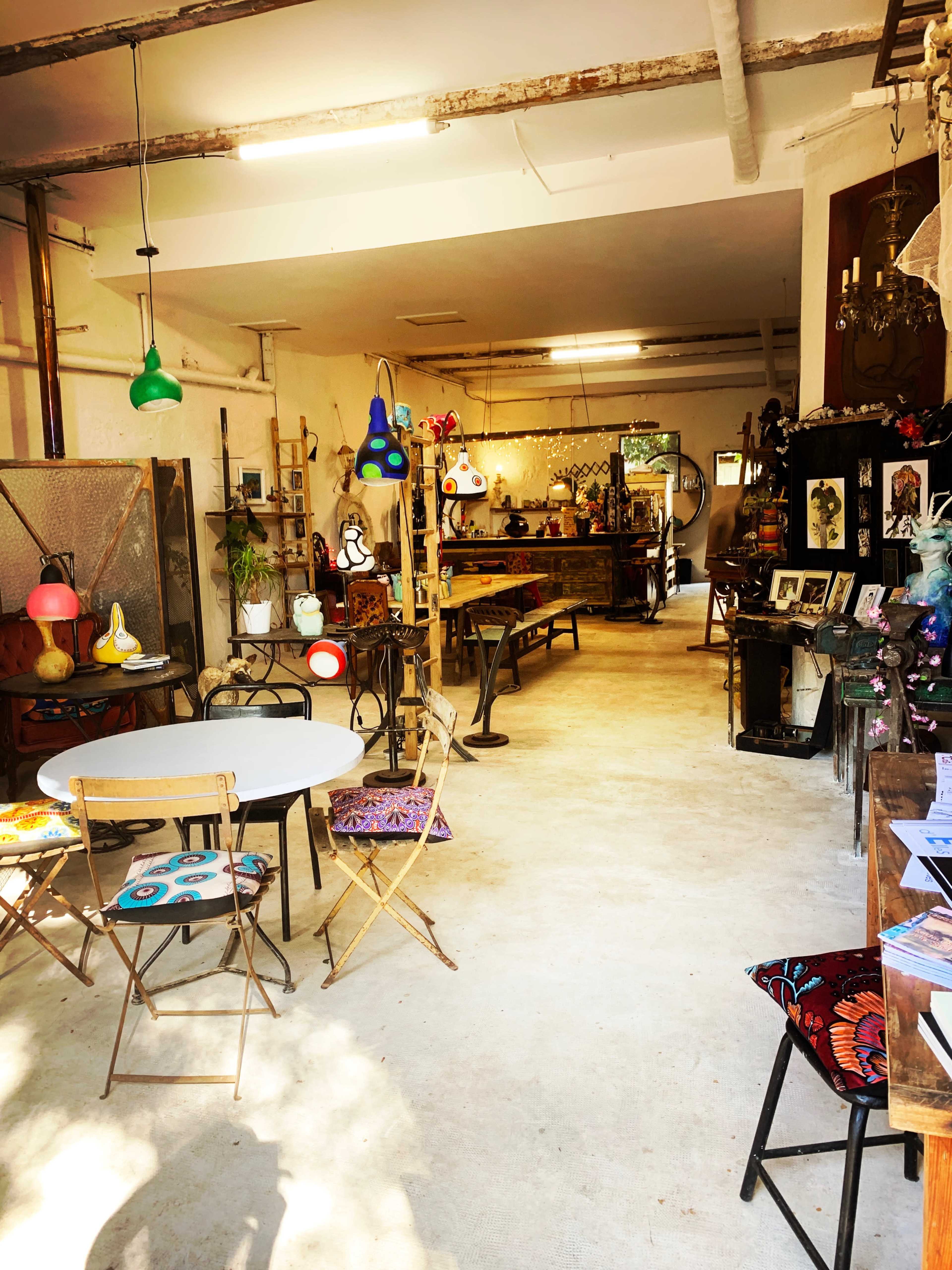The image shows an interior of a shop filled with various vintage items, furniture, and decorative objects, with a table and chairs set up for seating.