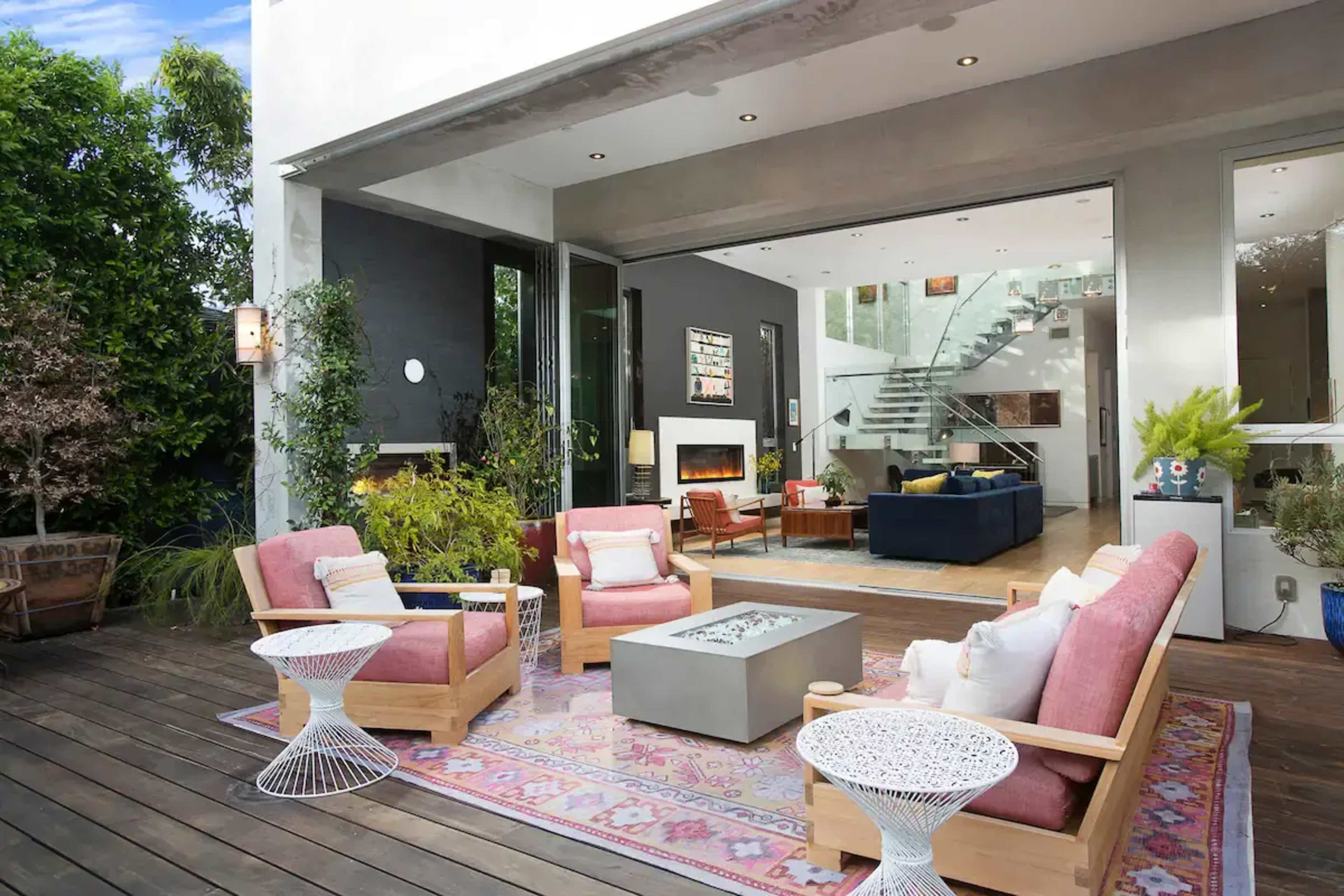 A modern outdoor living space featuring a patio with wooden flooring, pink chairs, a decorative rug, and a view of a glass-walled interior with a staircase.