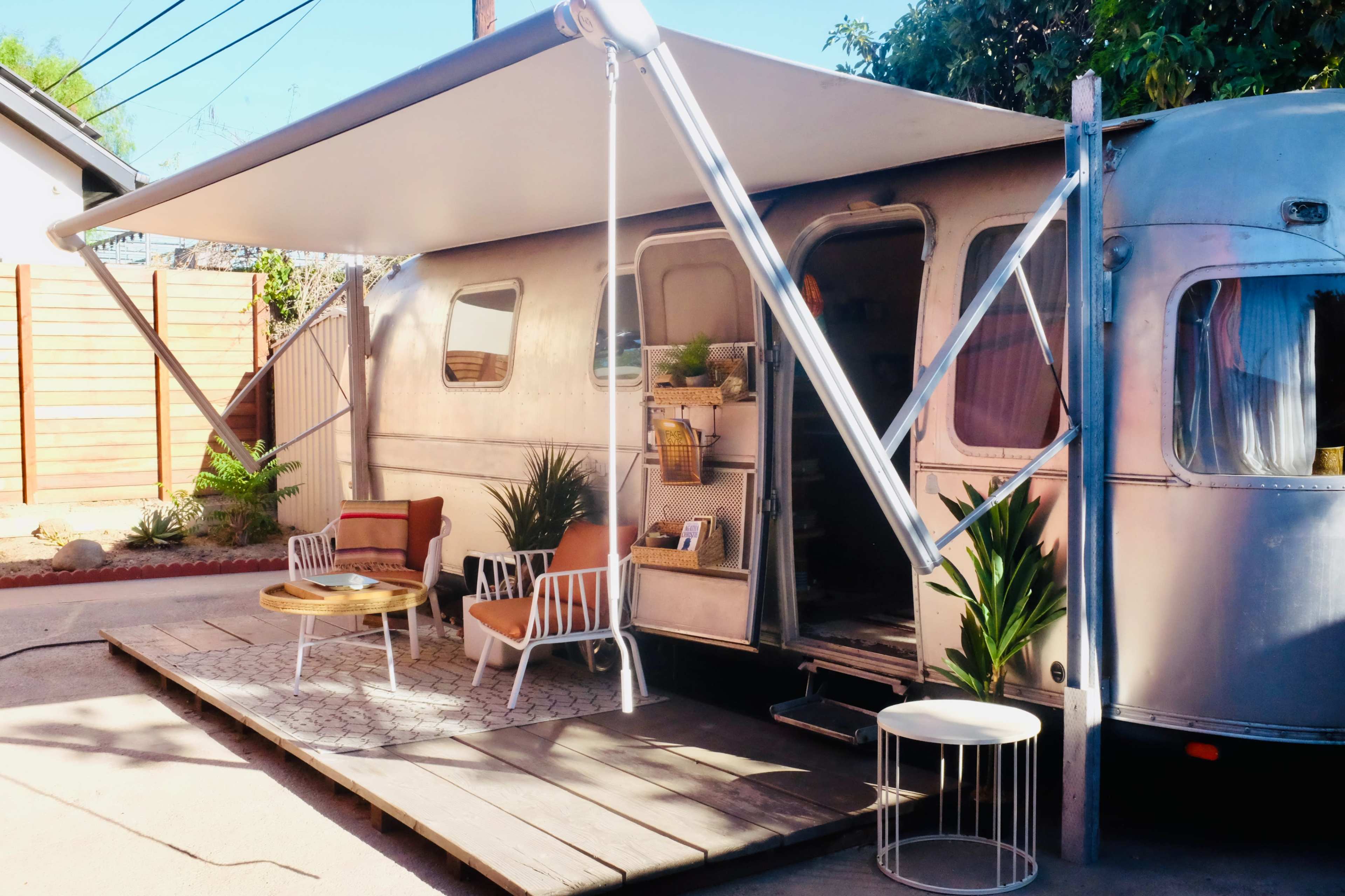A vintage Airstream trailer with a wooden deck, outdoor seating, and a large awning is positioned near a landscaped area.