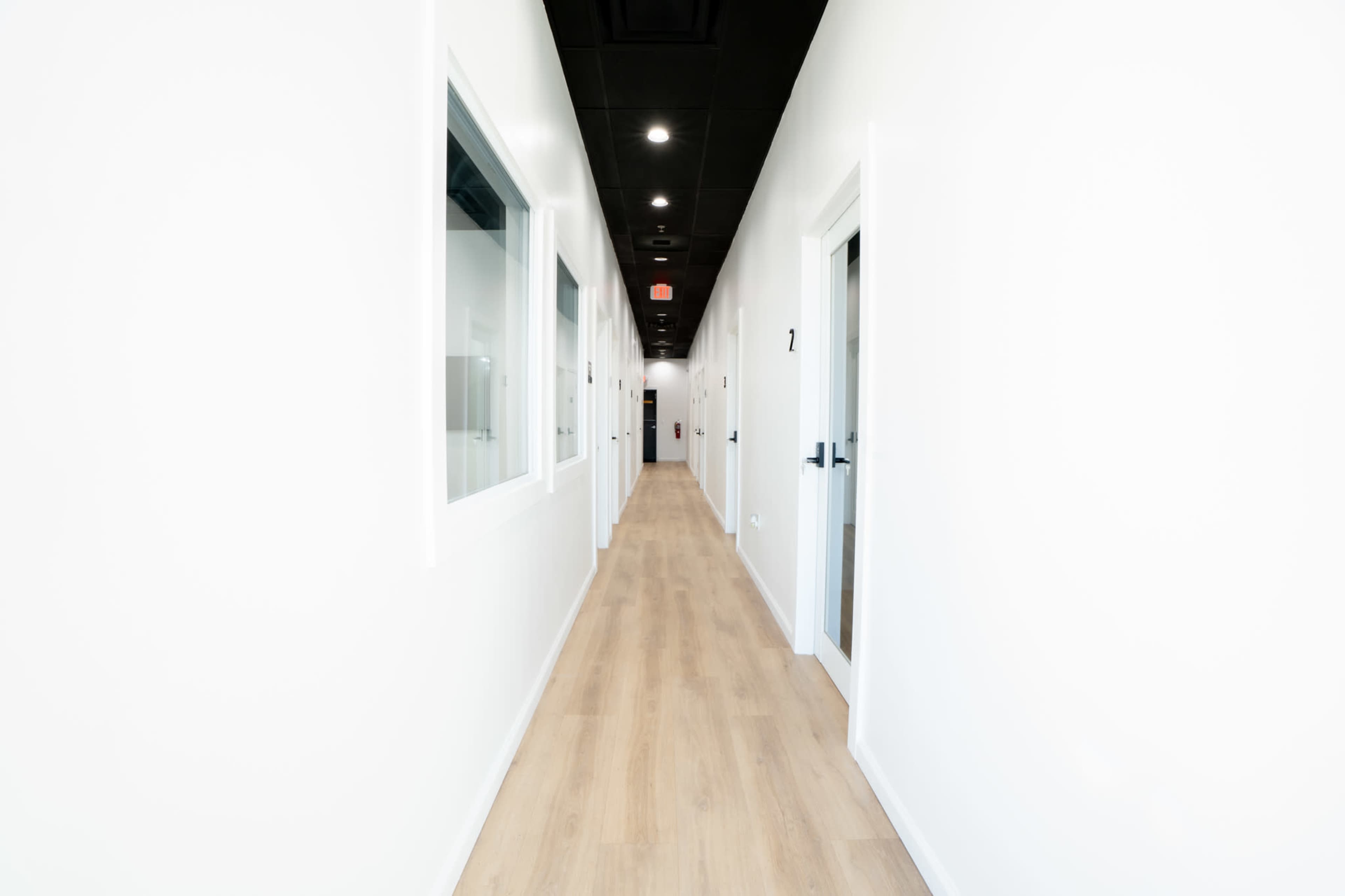 A long hallway with white walls and several doors on either side leads to a distant exit.