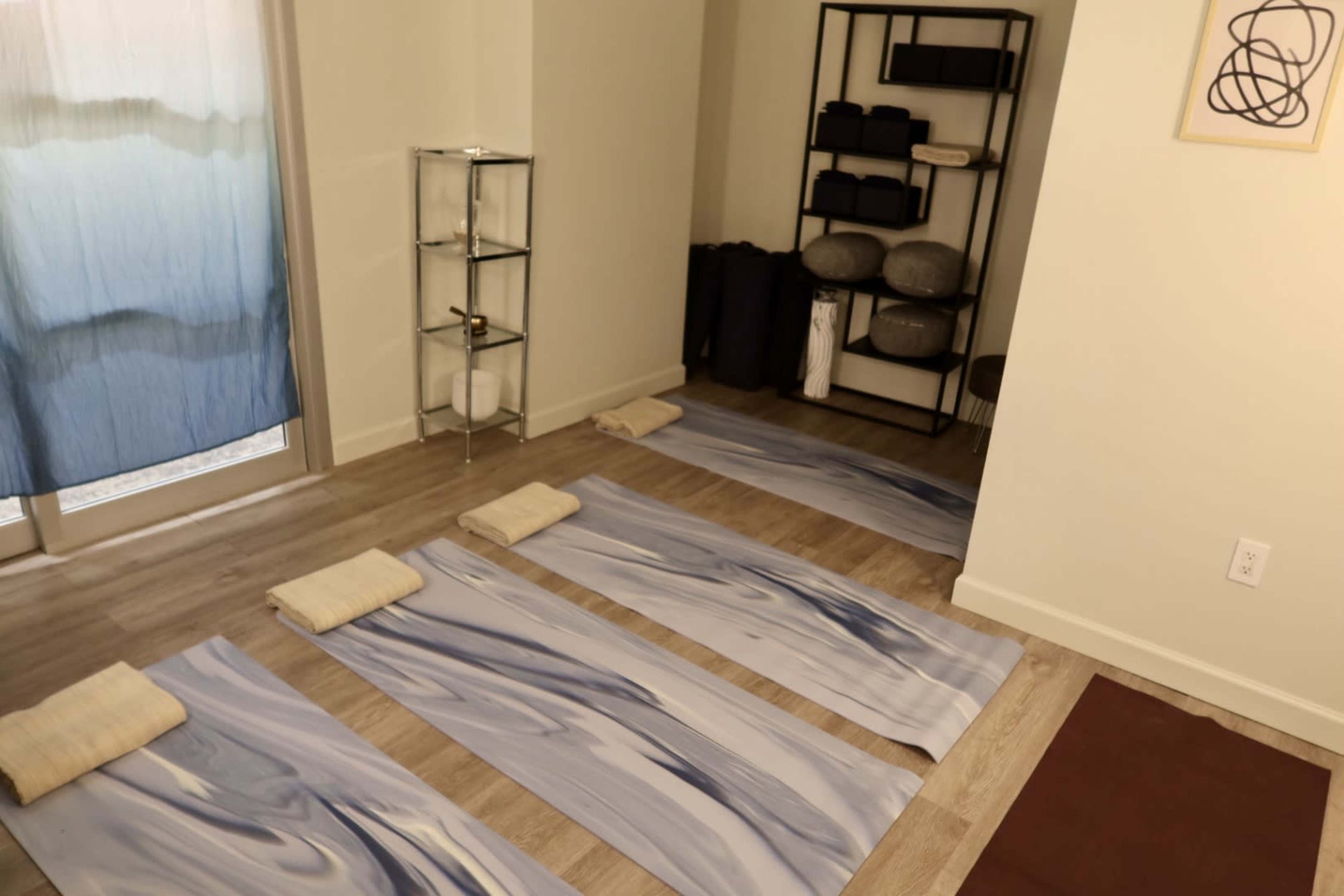 The image shows a minimalist yoga studio with four yoga mats placed on the wooden floor, a shelf storing yoga props, and large windows covered by a blue curtain.