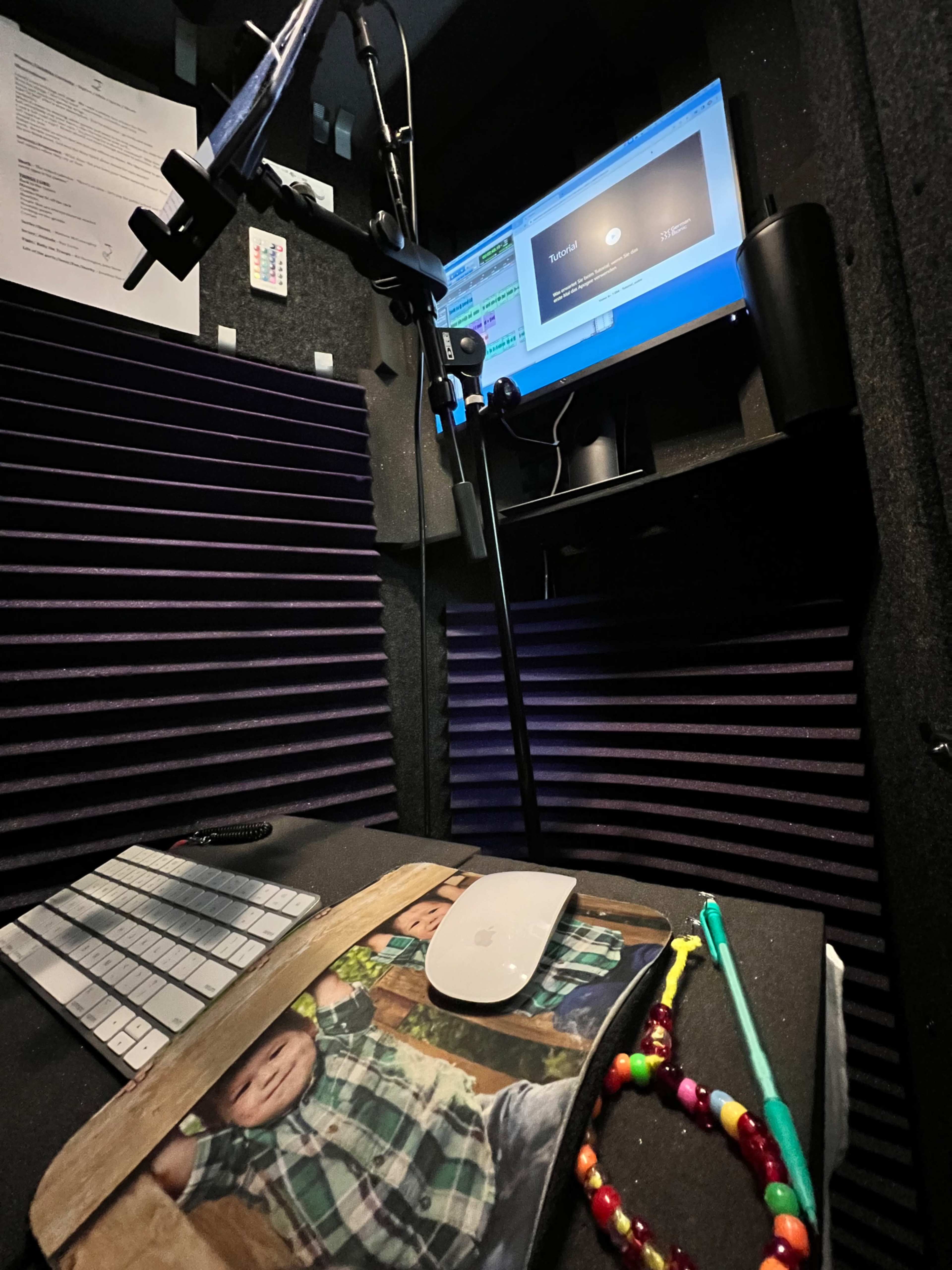 The image shows a recording booth with soundproofing panels, a computer monitor displaying software, a microphone on a stand, and various personal items on a desk.
