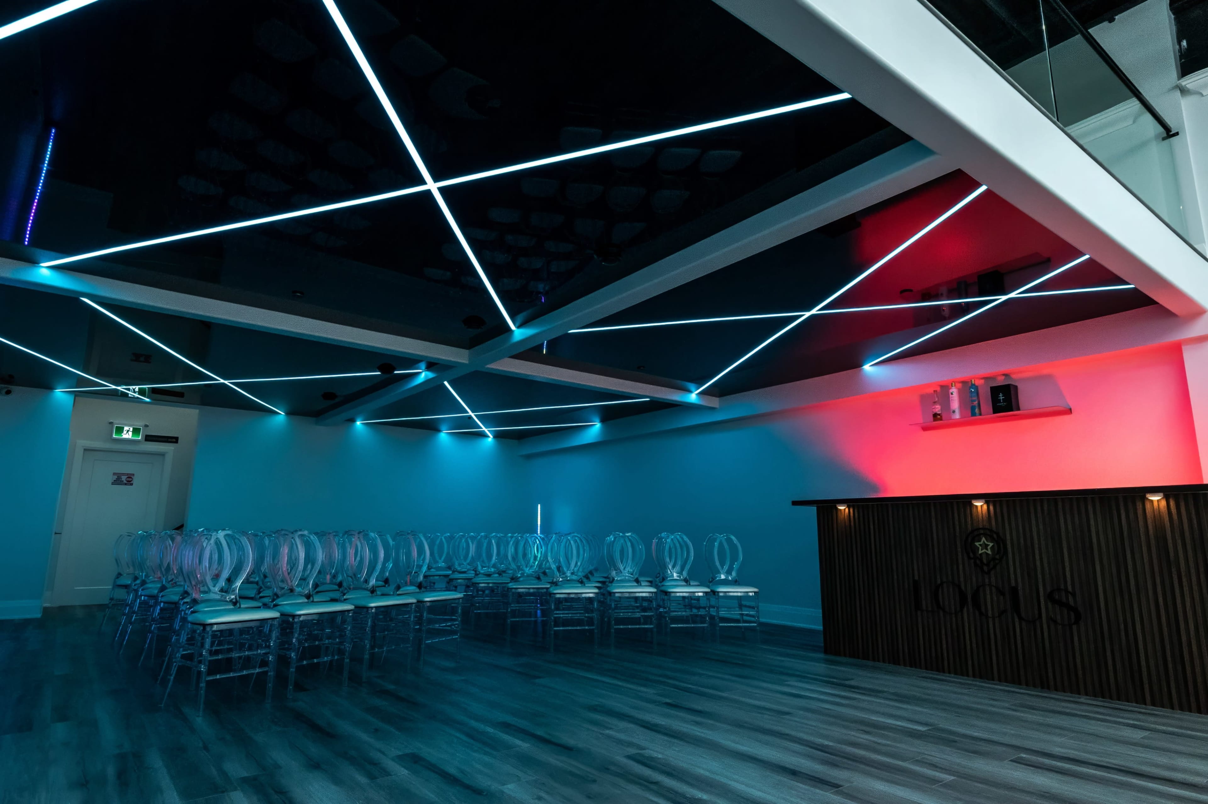 A modern event space with a mirrored ceiling and illuminated lines of blue and red light, featuring clear chairs arranged in rows facing a wooden bar marked "LOCUS."
