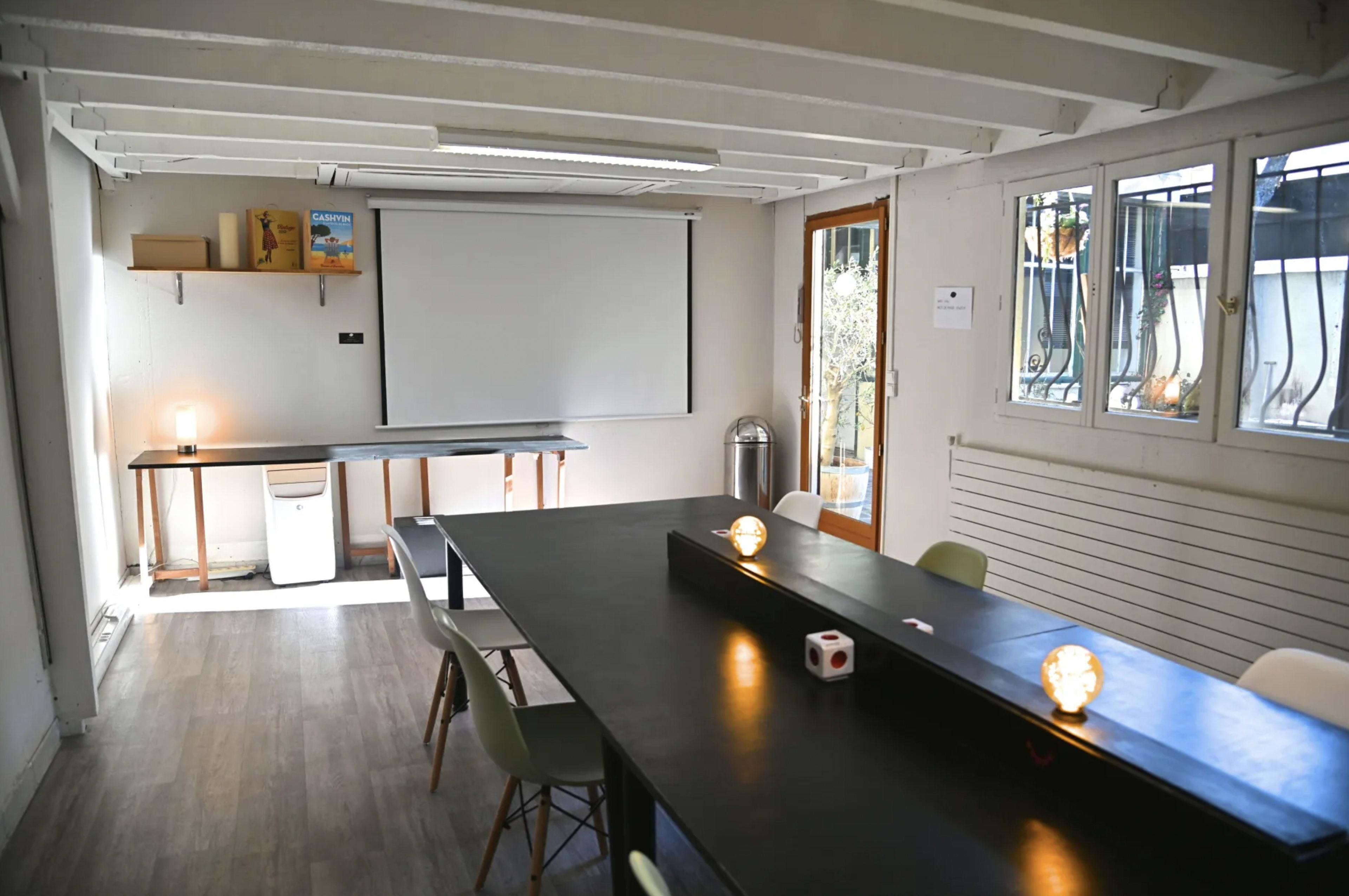 A minimalistic meeting room with a large black table, chairs, a projector screen, and a glass door leading to an outdoor area.