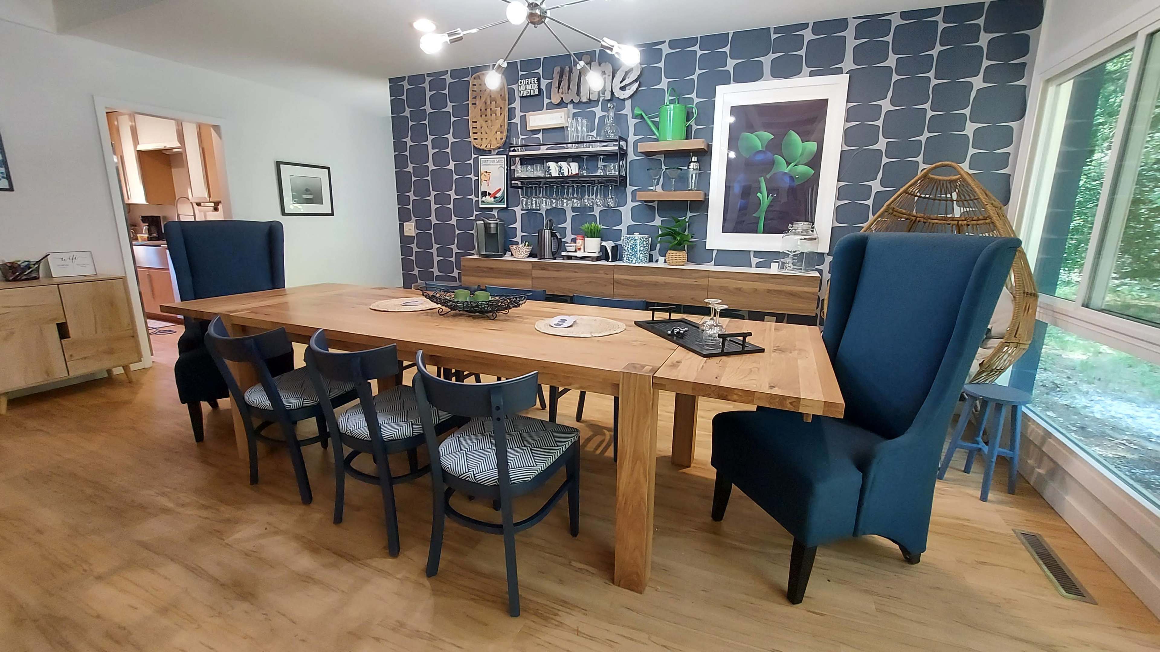 A spacious dining area with a large wooden table surrounded by high-backed chairs, set against a wall with blue patterned wallpaper and a wooden sideboard.