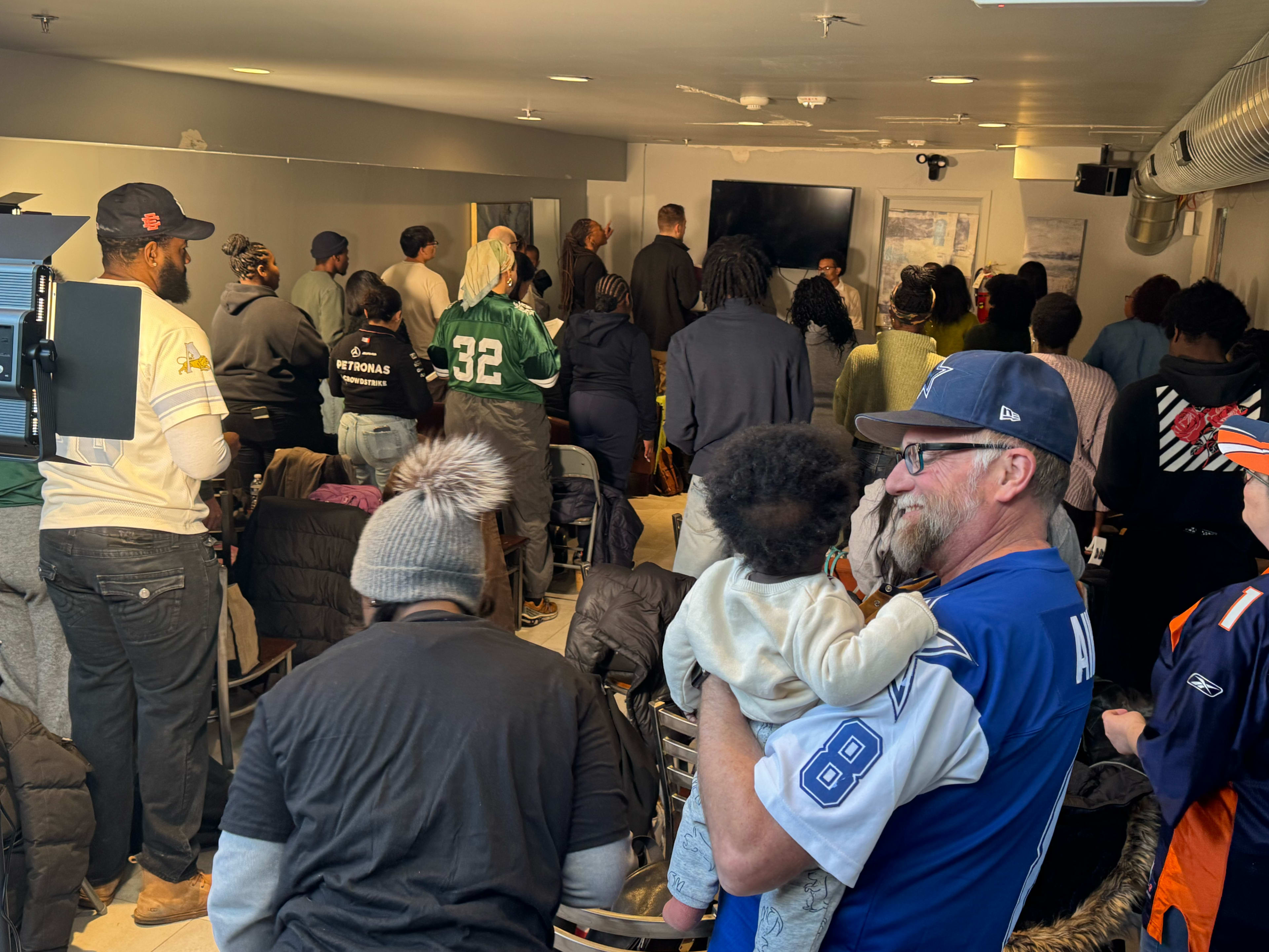 A diverse group of people stands in a crowded room, facing a presentation on a screen, while one man holds a child.