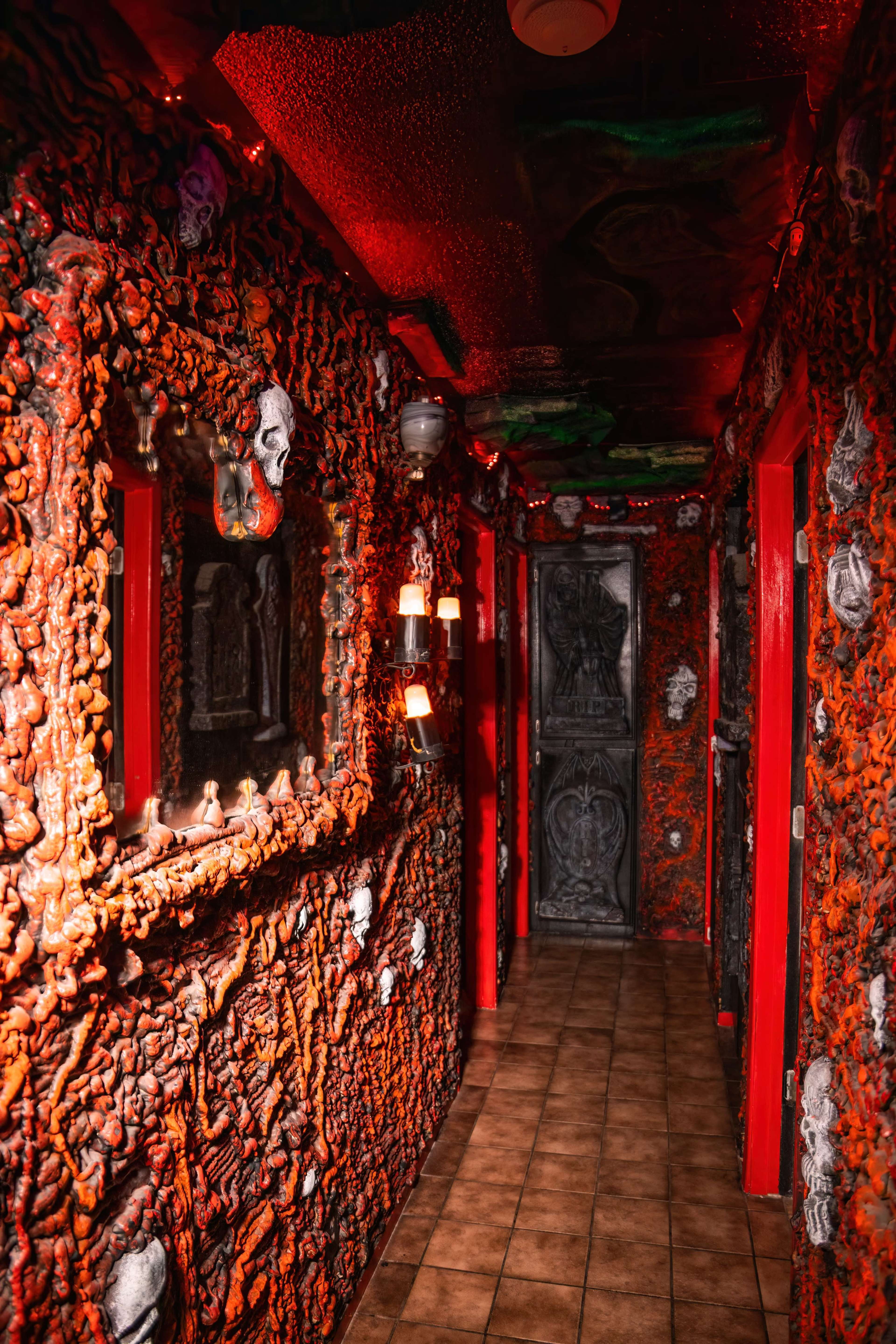 A dimly lit hallway features intricately textured orange walls adorned with skull and monster decorations, flanked by red doors and a dark, ornate door at the end.