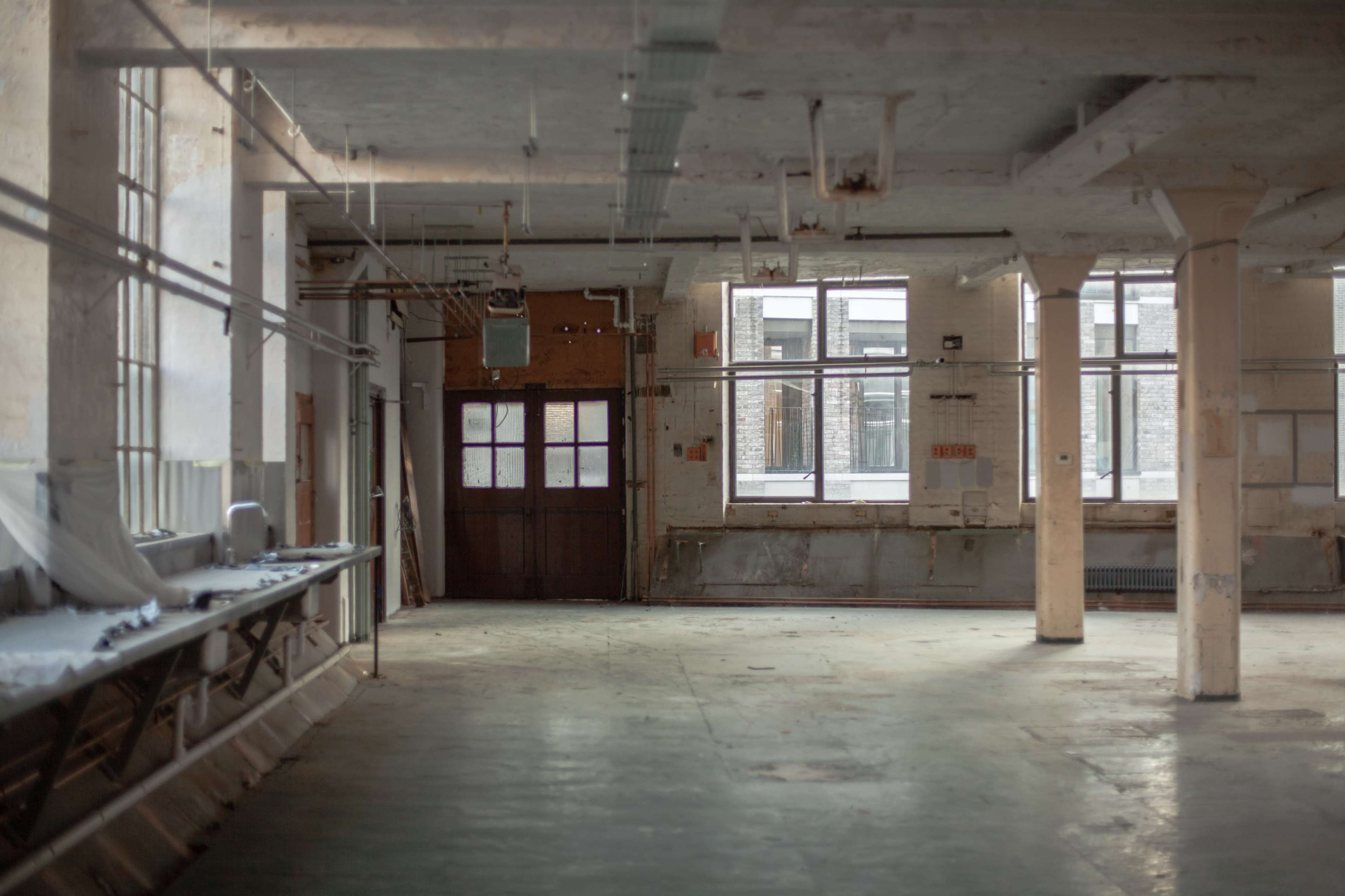 An empty industrial space features large windows and exposed columns, with a concrete floor and minimal furnishings.
