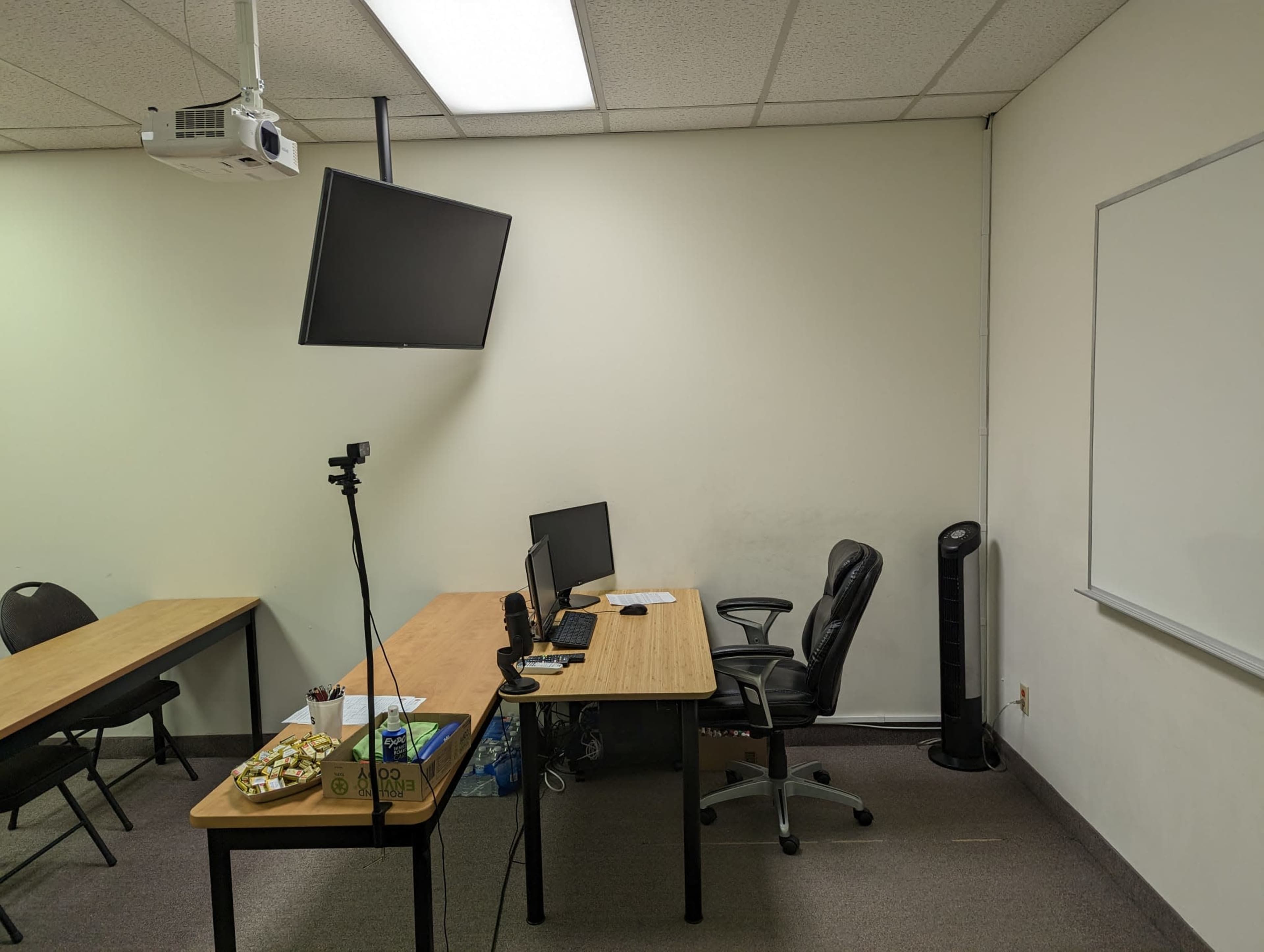 A classroom with two tables, an empty office chair, a computer setup, a projector mounted on the ceiling, and a whiteboard on the wall.