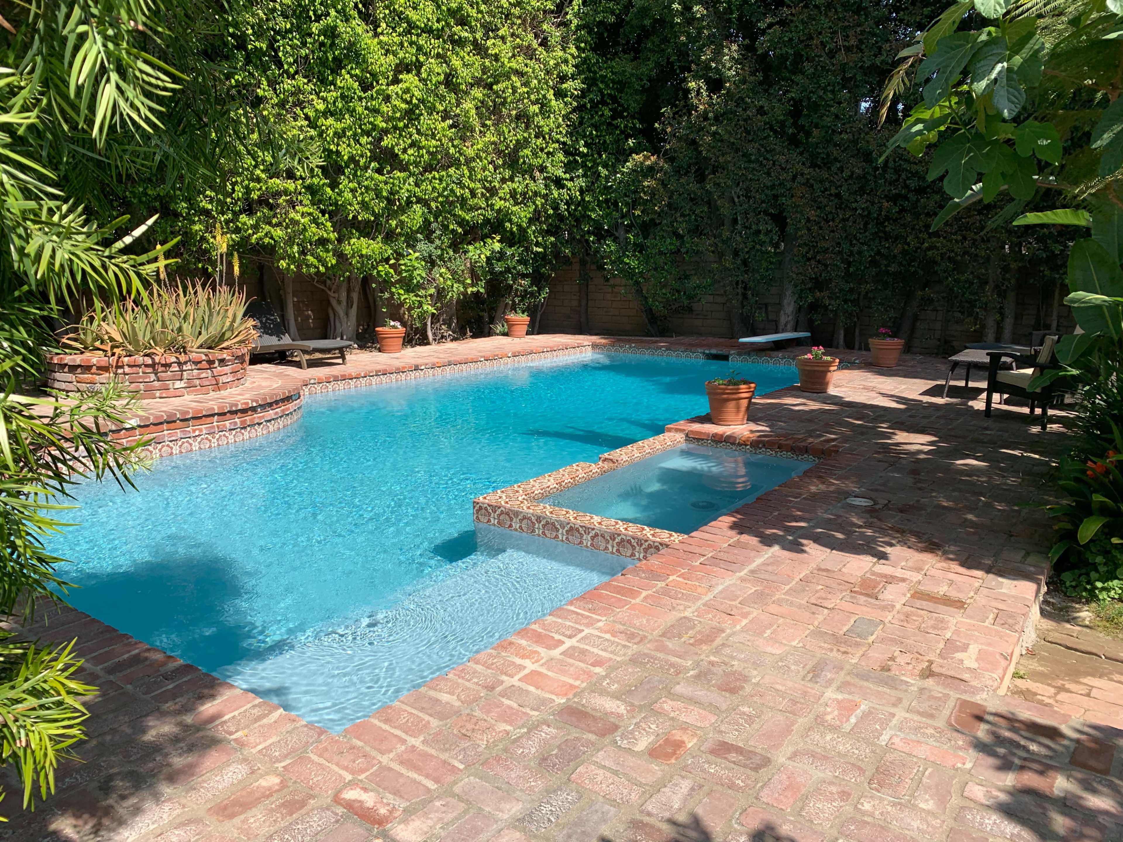 A brick patio surrounding a clear blue swimming pool, bordered by lush greenery and terracotta planters.