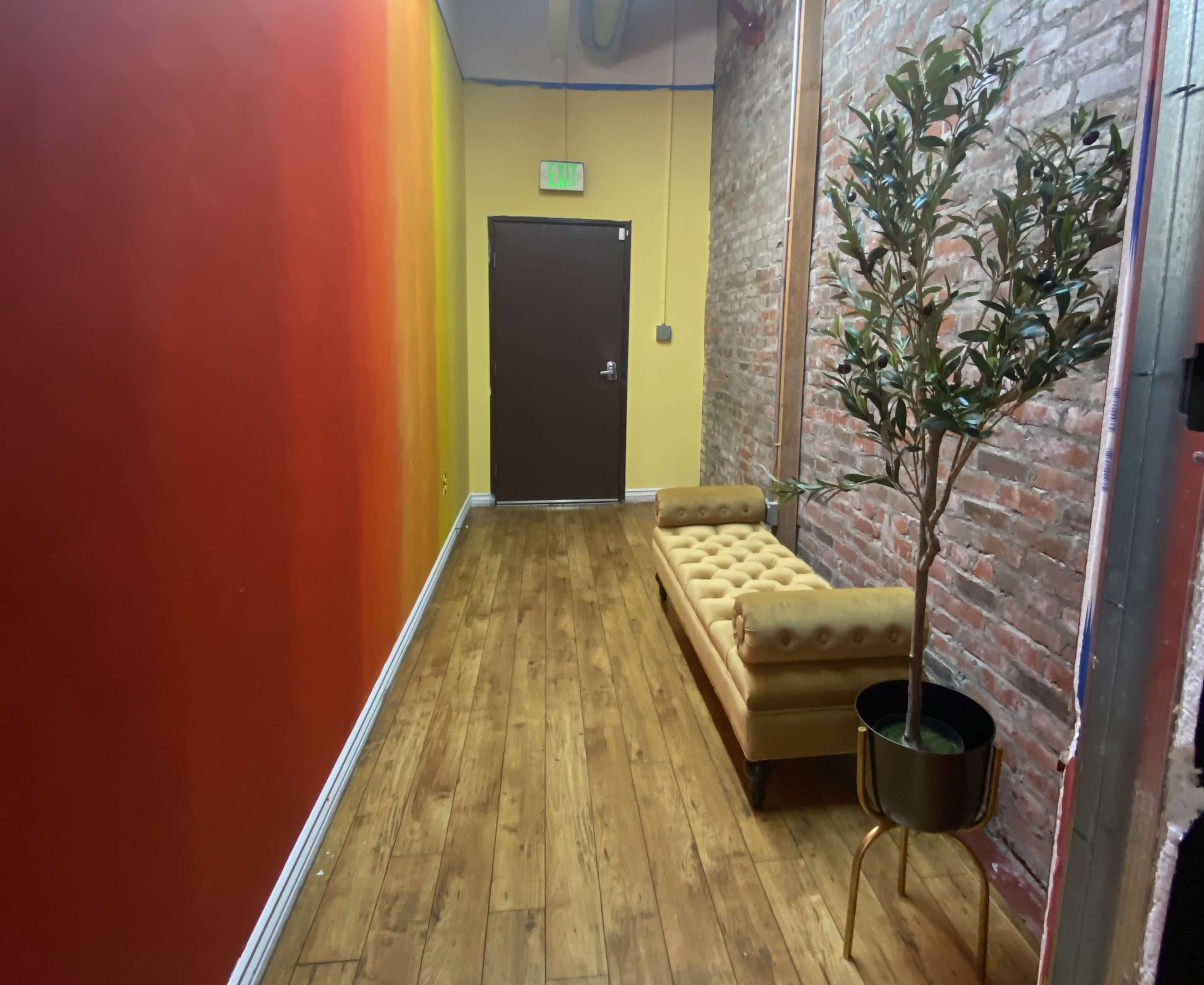 A narrow hallway features a tufted yellow couch against a brick wall, with a potted plant and contrasting painted walls in shades of red and orange.