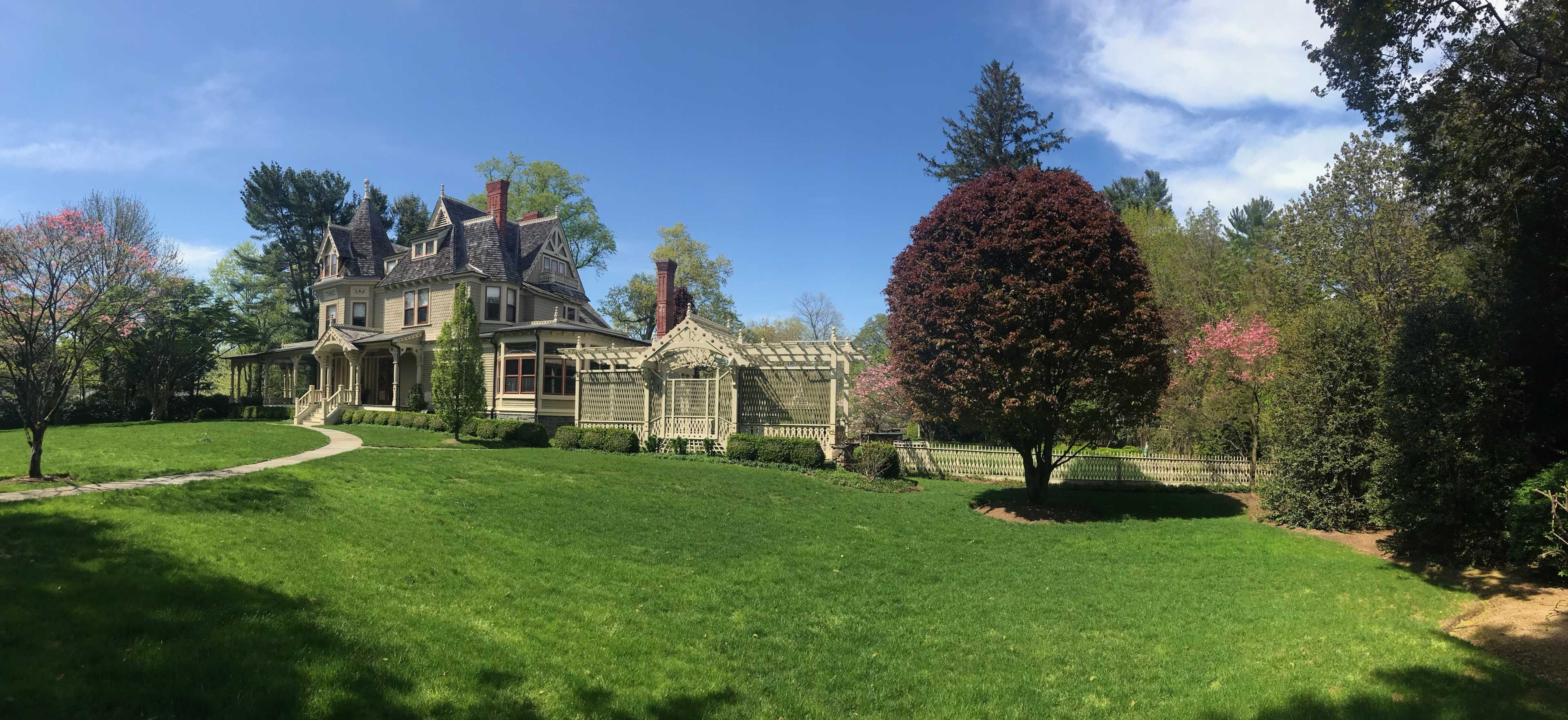 A large Victorian-style house with intricate architectural details stands beside a manicured lawn and colorful trees under a clear blue sky.